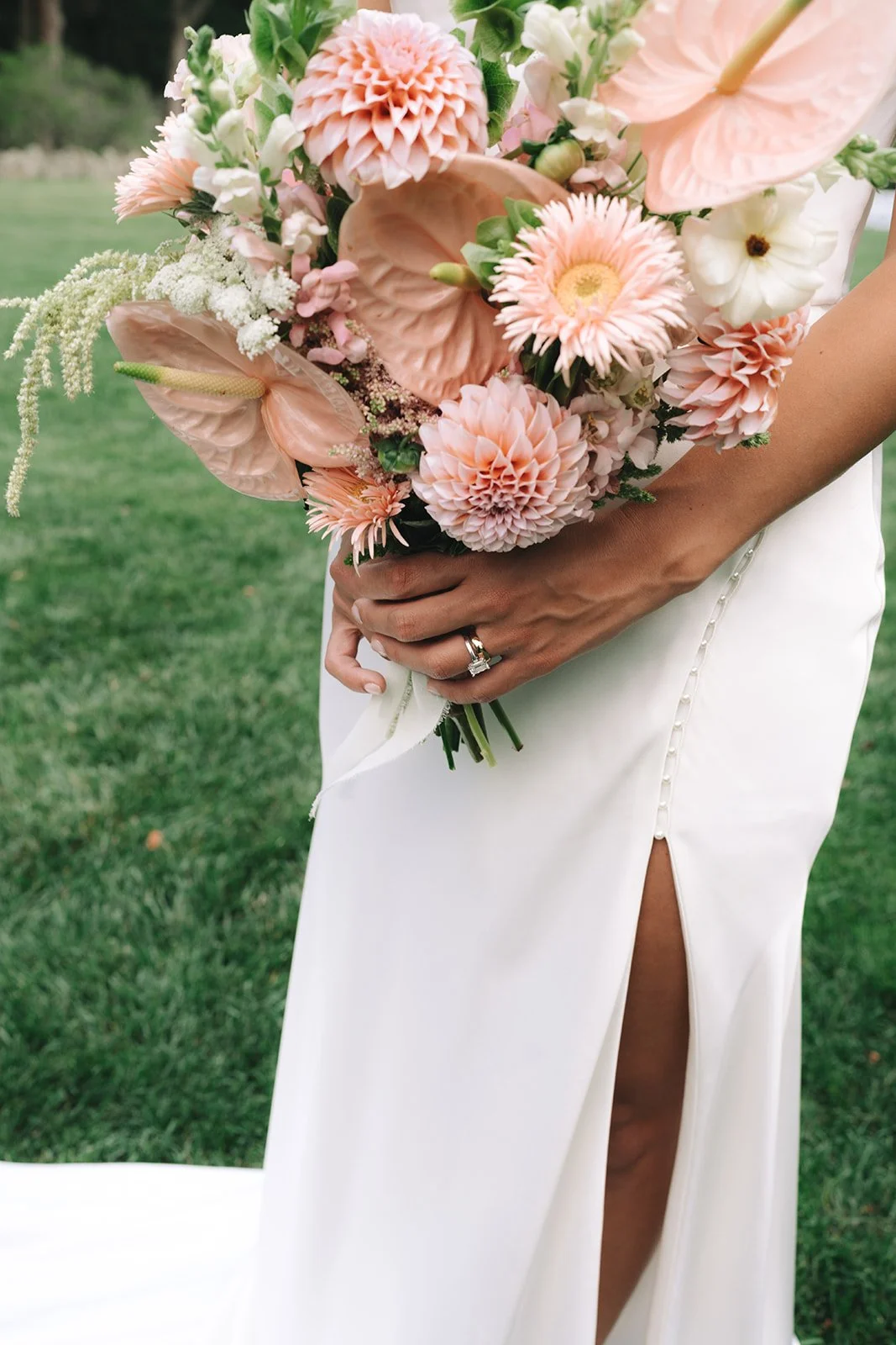 A woman in a white dress holding a bouquet of pink and white flowers outdoors, showing her hand with a wedding ring and a slit in her dress revealing her leg.