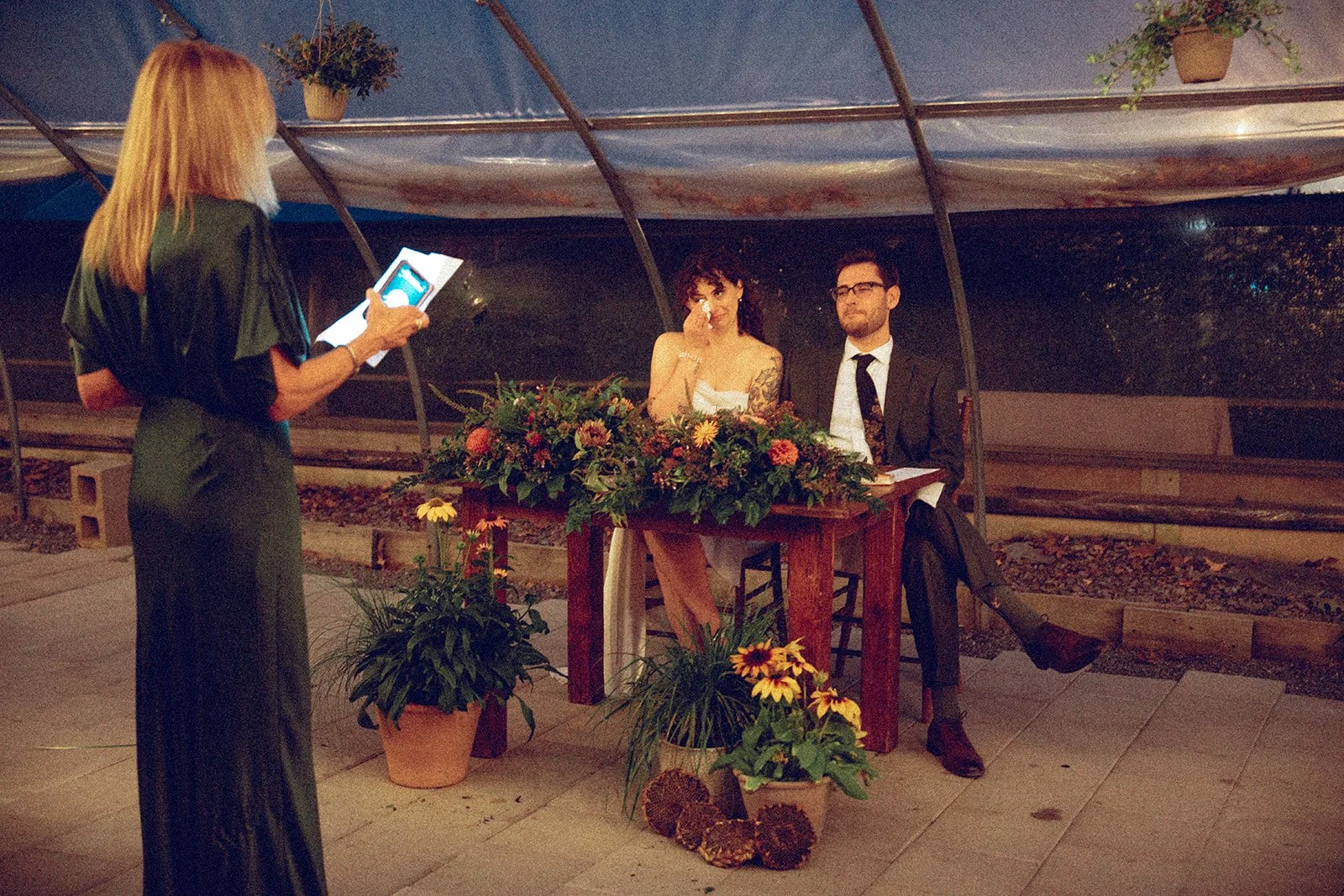 Two people, a woman with tattoos and a man in a suit, sitting at a table with flowers, being filmed or photographed by a woman with a tablet in an outdoor setting under a canopy