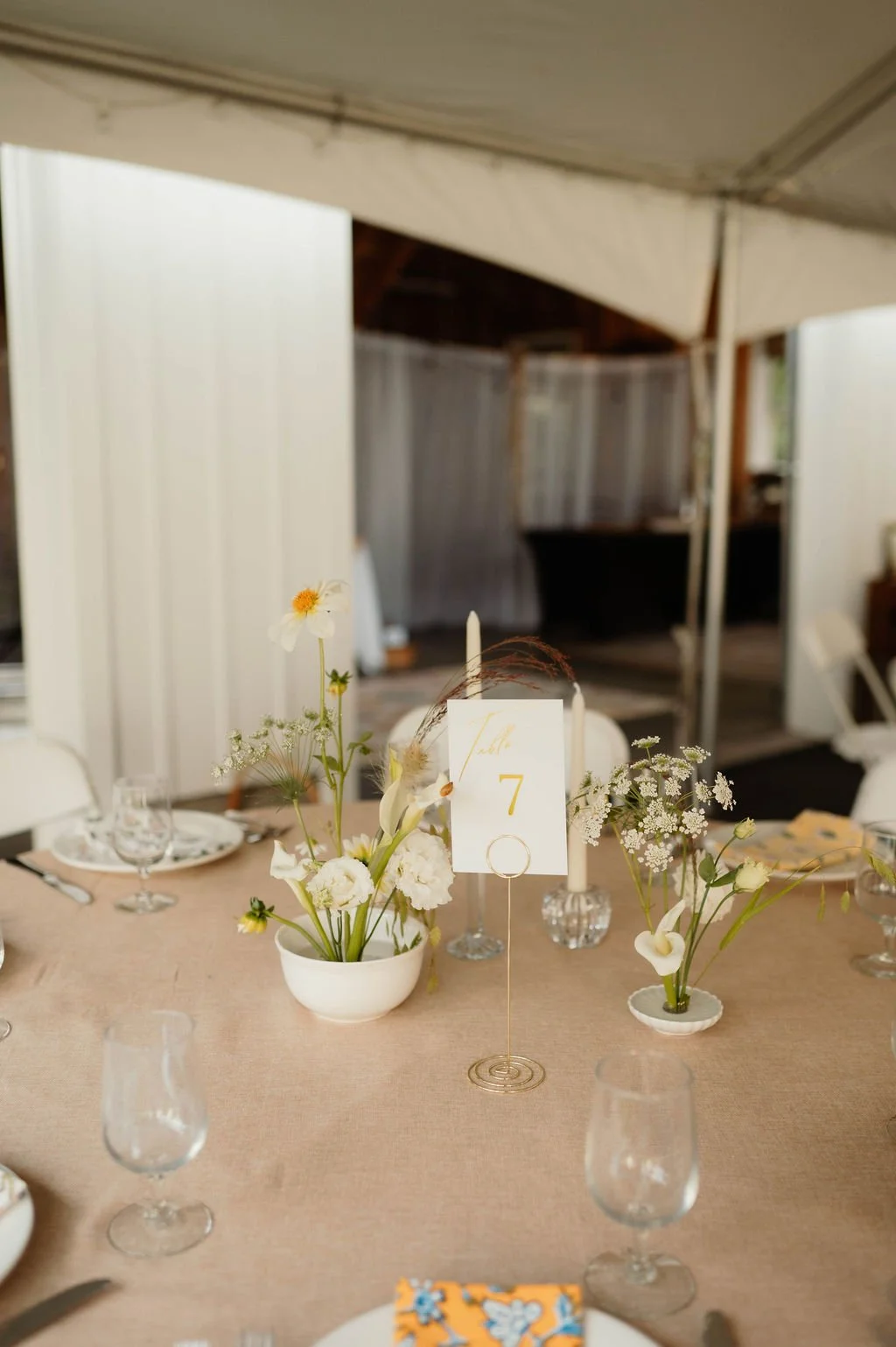 A table setting at a wedding reception with a floral centerpiece in white bowls, candles, and a table number card with the number 7. There are wine glasses, plates, and cutlery on the table, with a rustic indoor venue in the background.