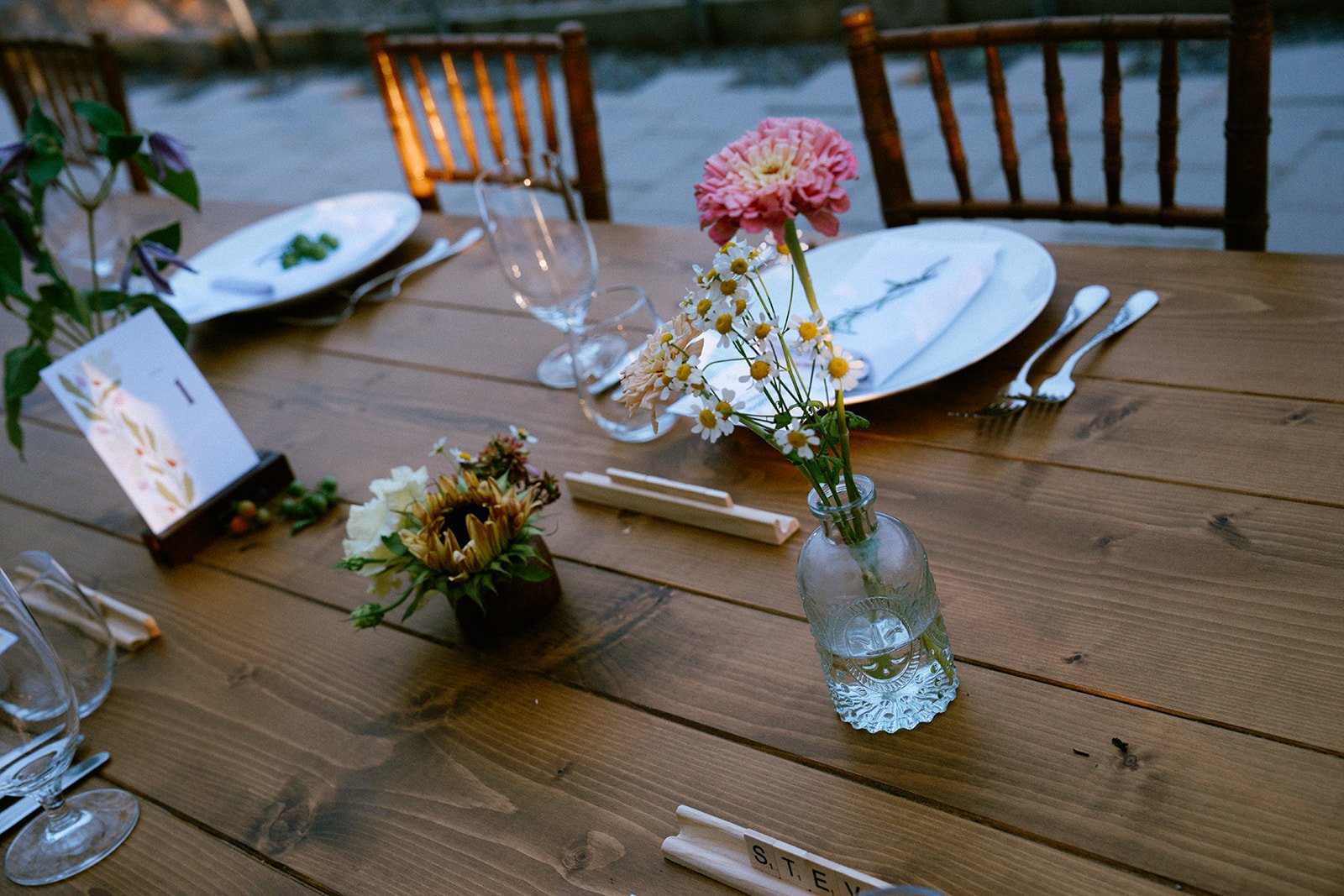 A wooden outdoor dining table set for a meal with floral centerpieces, empty plates, glassware, cutlery, and a table number card.