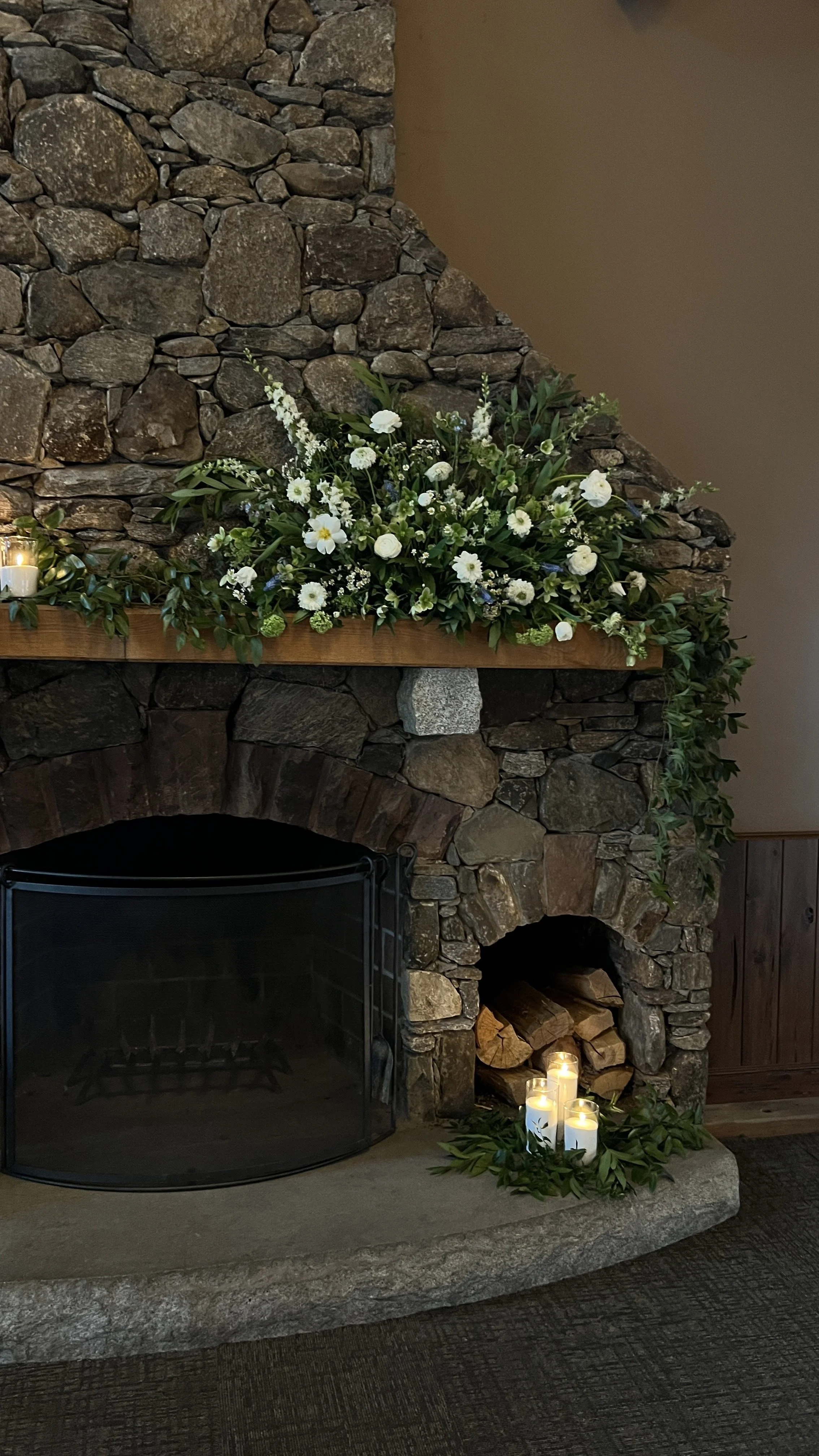 A stone fireplace decorated with white flowers, candles, and greenery for a cozy setting.