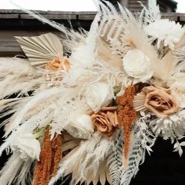 A floral arrangement featuring white roses, beige roses, dried grasses, and palm leaves.