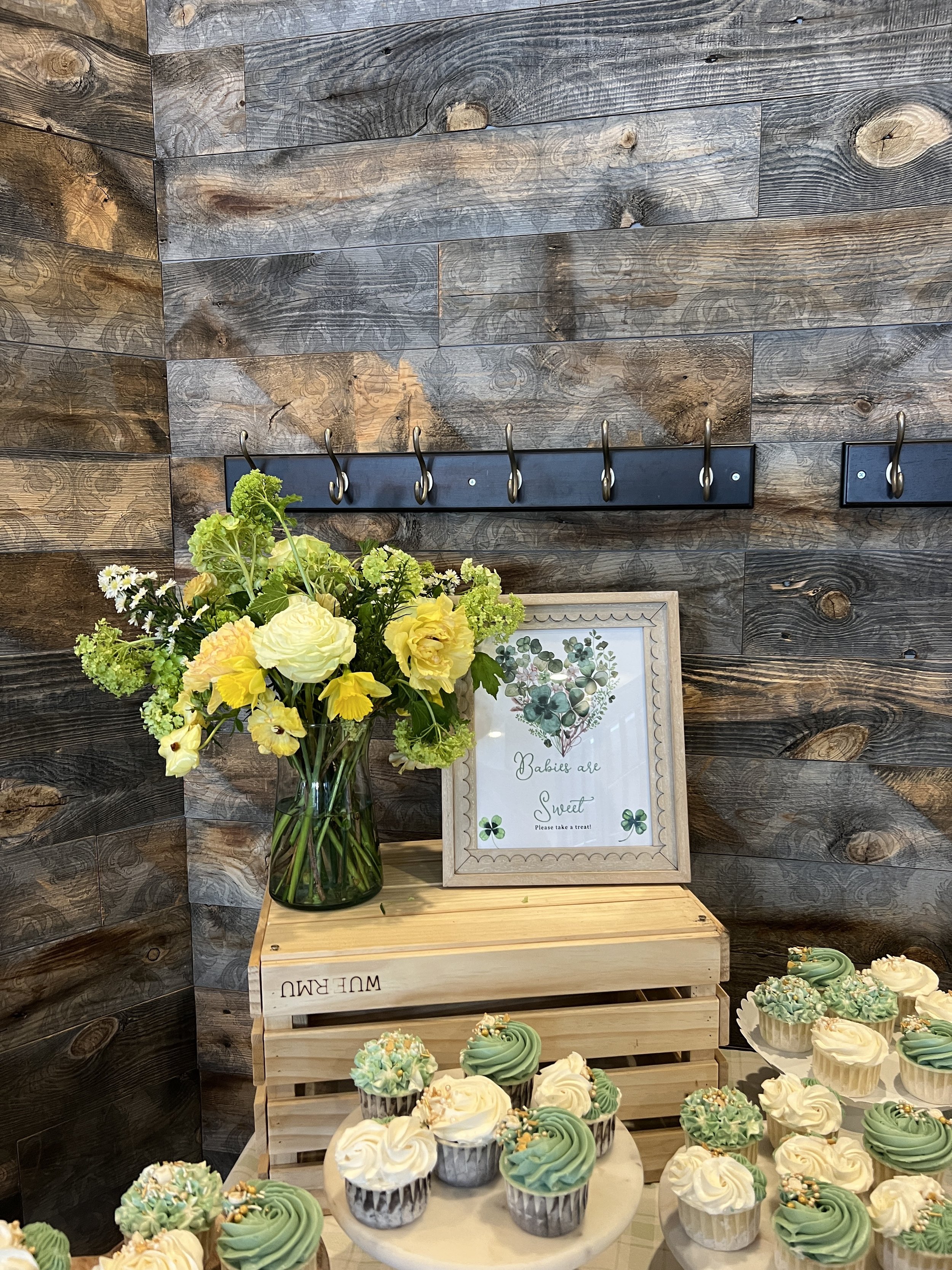 A display of cupcakes with green and white frosting, decorated with gold sprinkles, on a table with a wooden crate, a framed sign, and a vase of yellow flowers against a dark wooden wall with hooks.