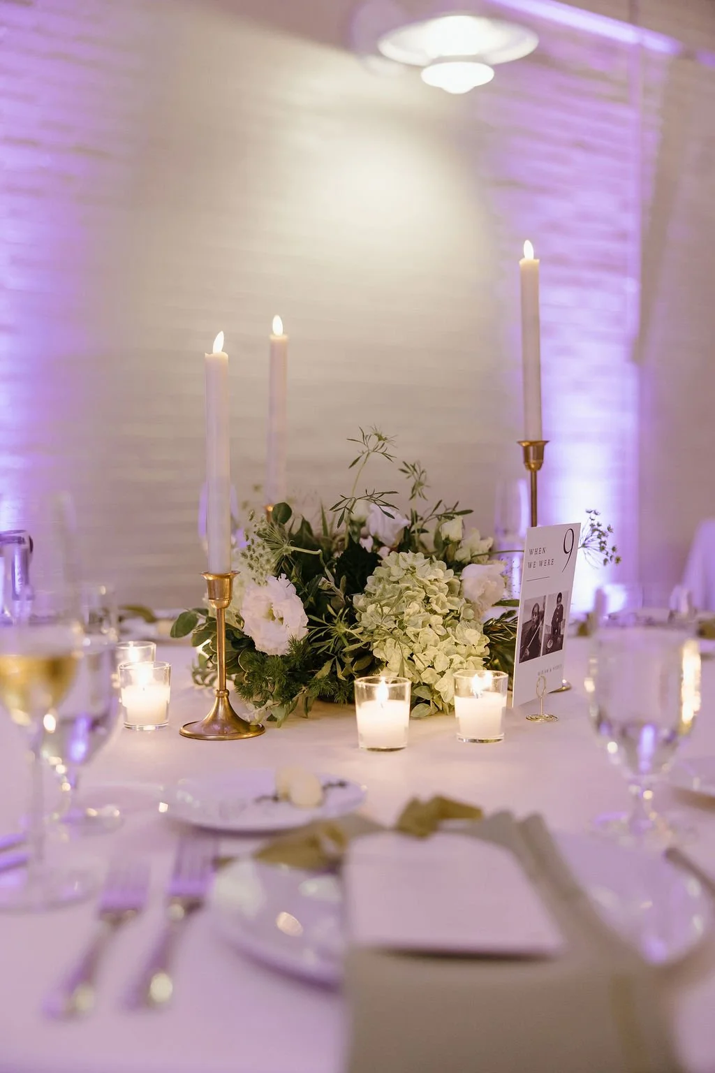 A decorated wedding reception table with white flowers, candles, and tall white taper candles in gold holders, set against a softly lit wall with purple uplighting.