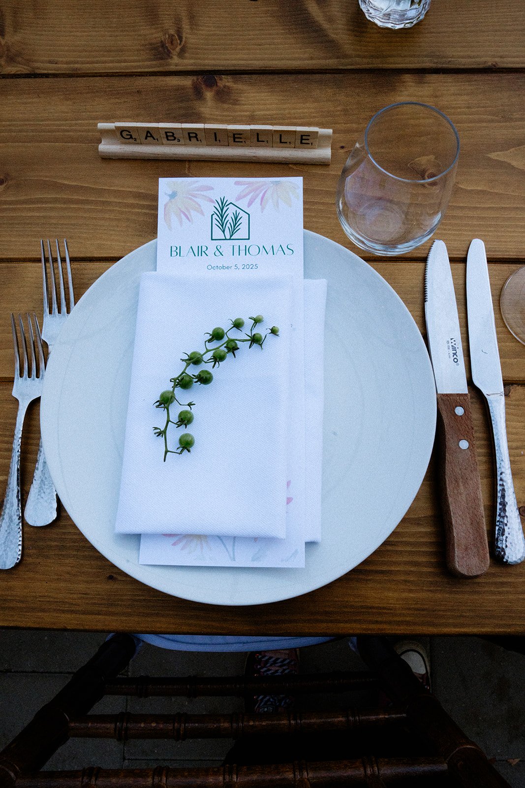 Elegant table setting with white plate, silverware, white napkin with green berries, wedding program for Blair & Thomas, and a water glass on a wooden table. Name 'Gabrielle' spelled out in wooden Scrabble tiles at the top.