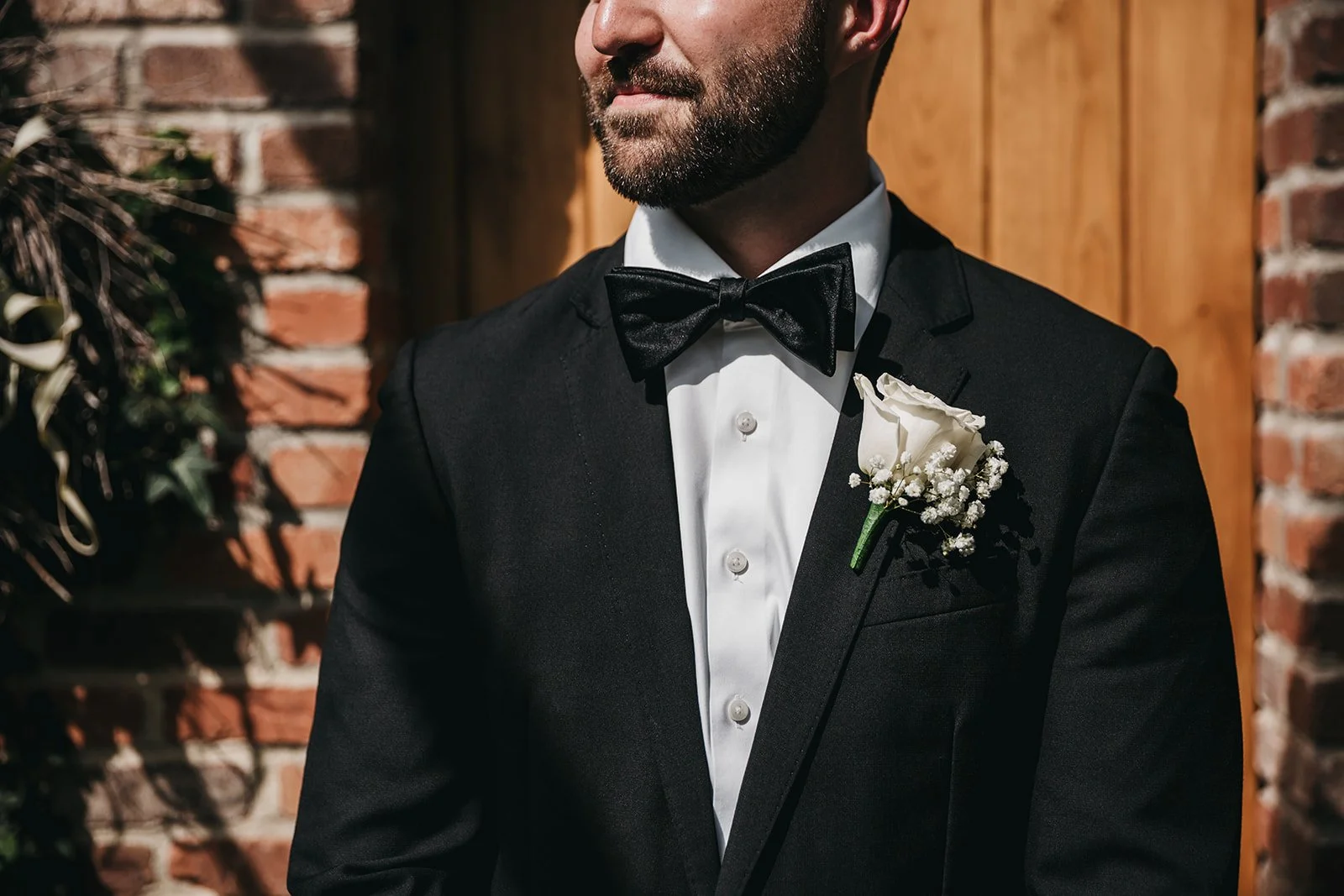 Man wearing a black tuxedo with a white shirt, black bow tie, and a white boutonniere, standing outdoors against a brick wall and wooden background.