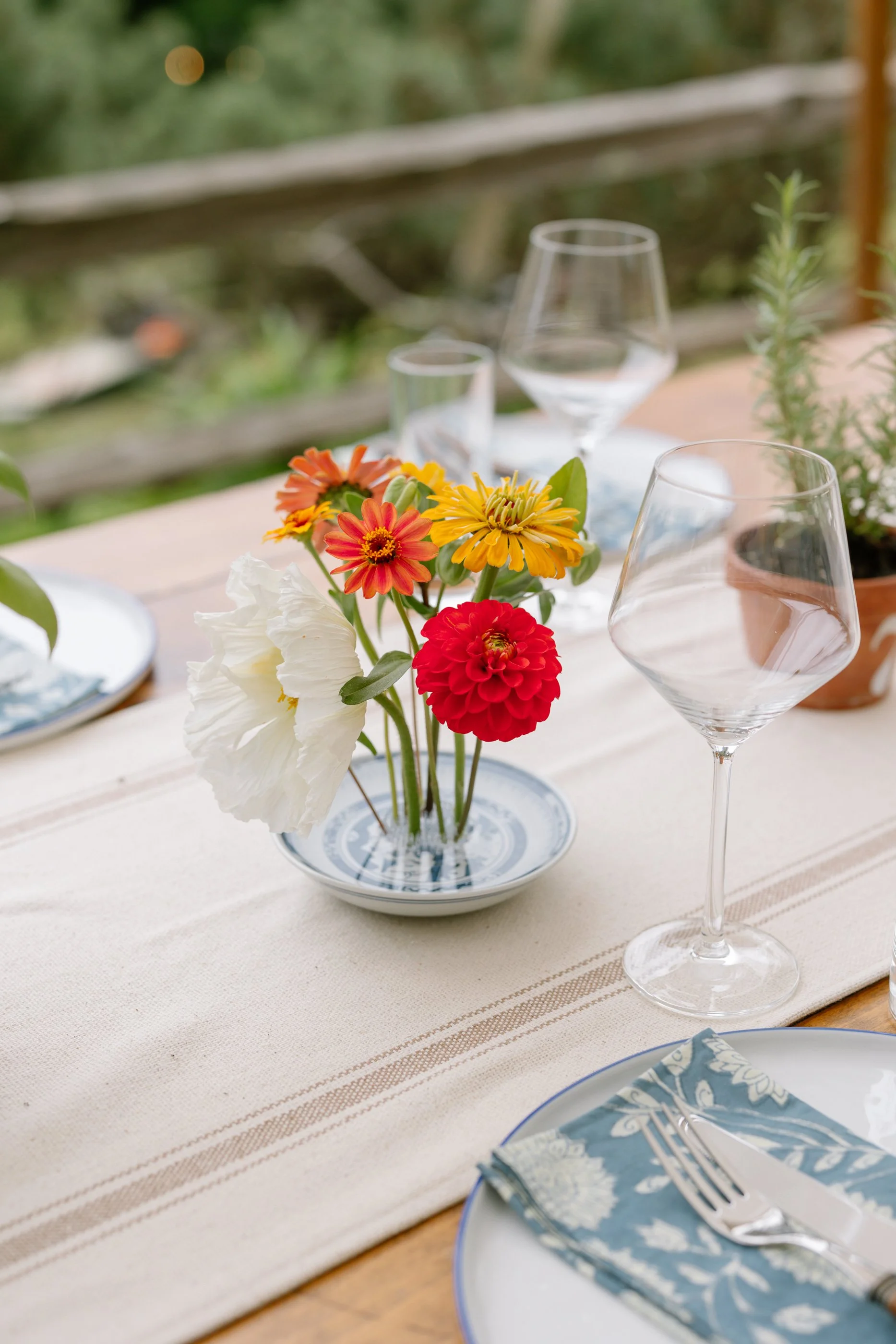 A table setting with a vase of colorful flowers, empty wine glasses, a plate with a napkin, and potted plants outdoors.