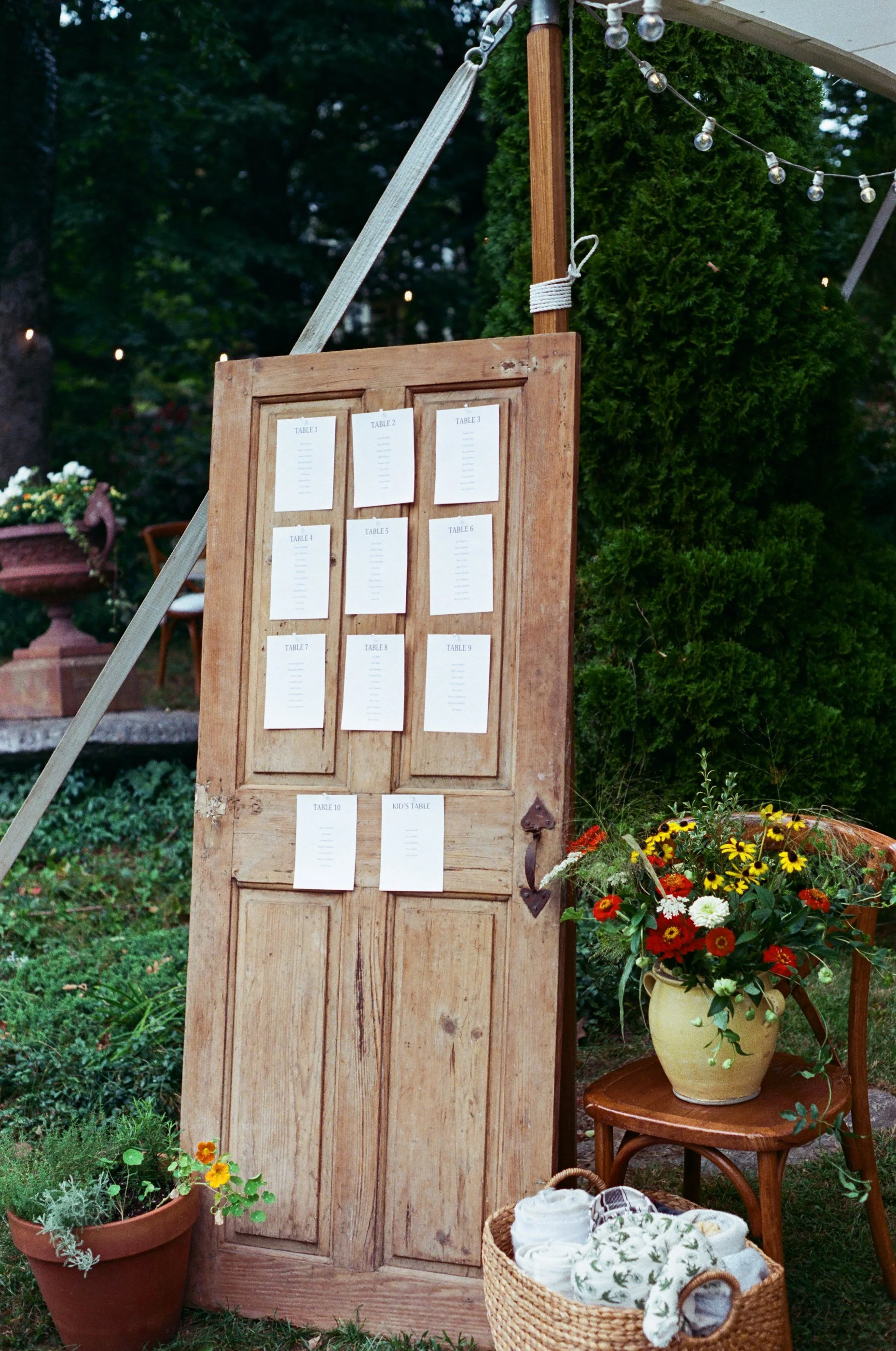 A rustic wooden door with seating cards attached, set up outdoors with potted plants, flowers, and string lights for a gathering or event.
