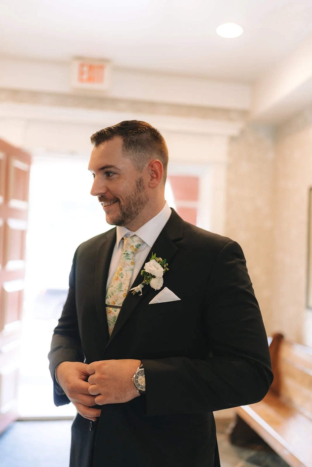 Man dressed in black suit with a white shirt and floral tie, adjusting his suit jacket inside a well-lit building.