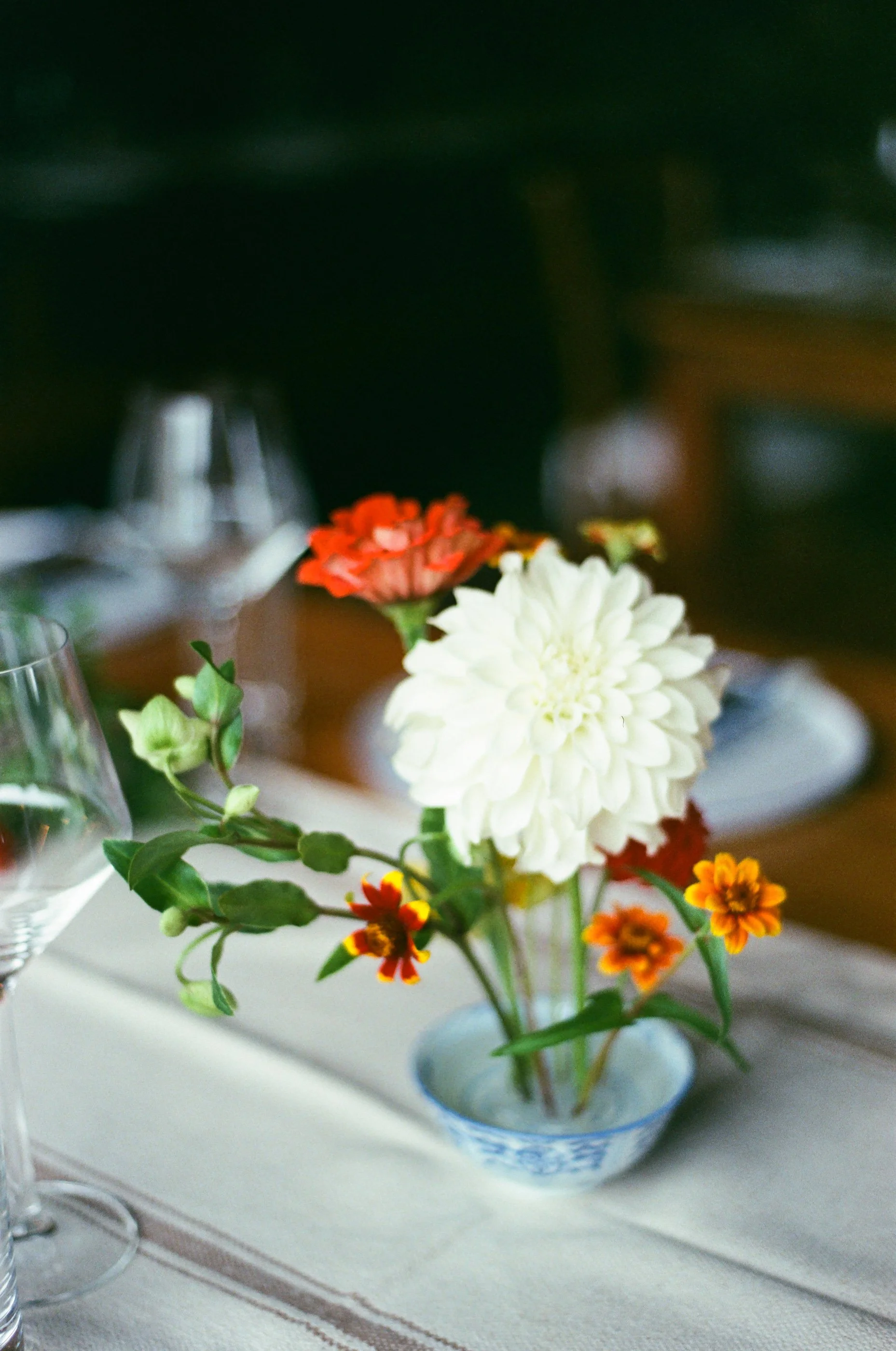 A small floral centerpiece with white, orange, and red flowers in a blue and white patterned vase, placed on a white tablecloth on a dining table.