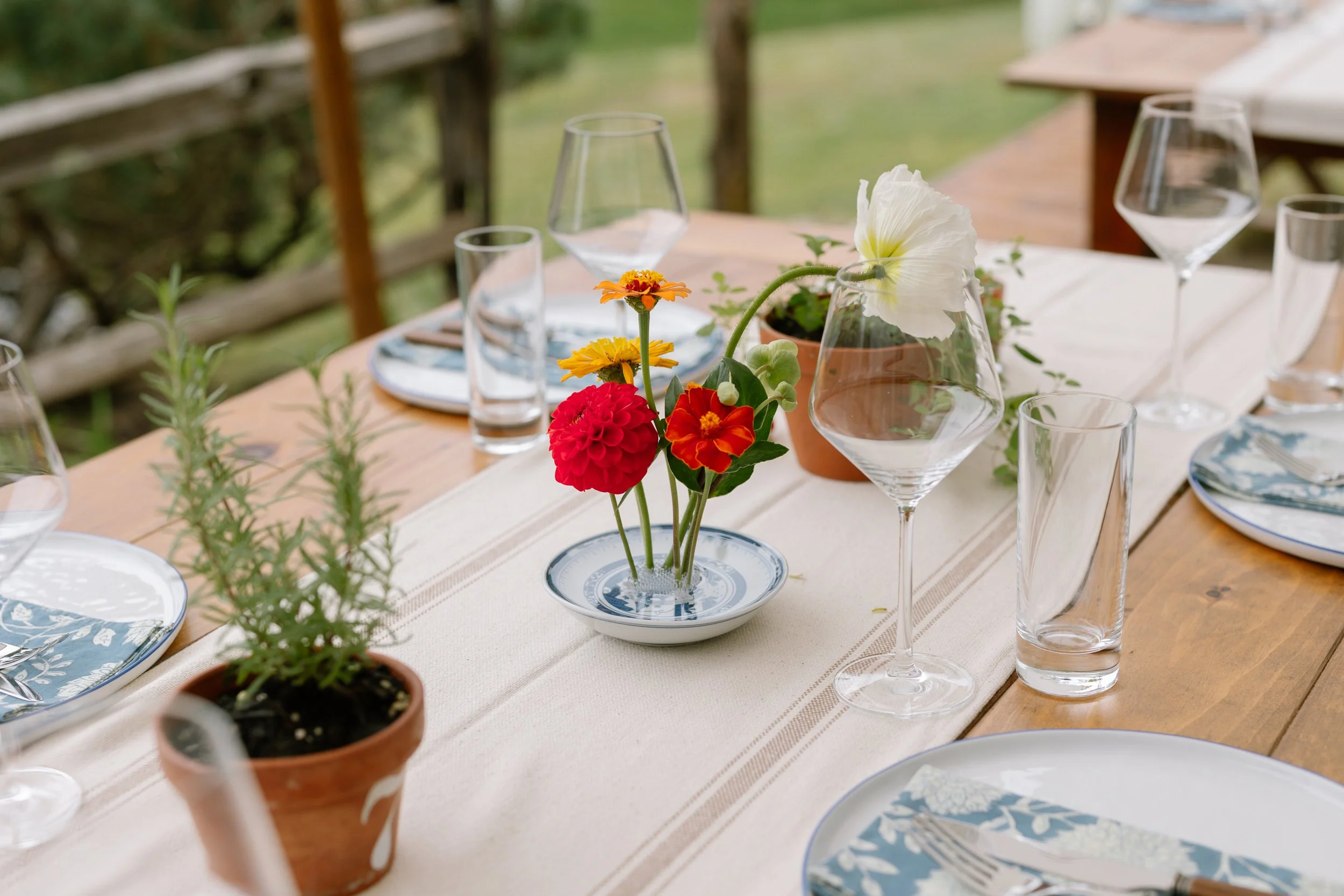 A table decorated with a vase of colorful flowers, glassware, plates with floral napkins, and potted plants outdoors.