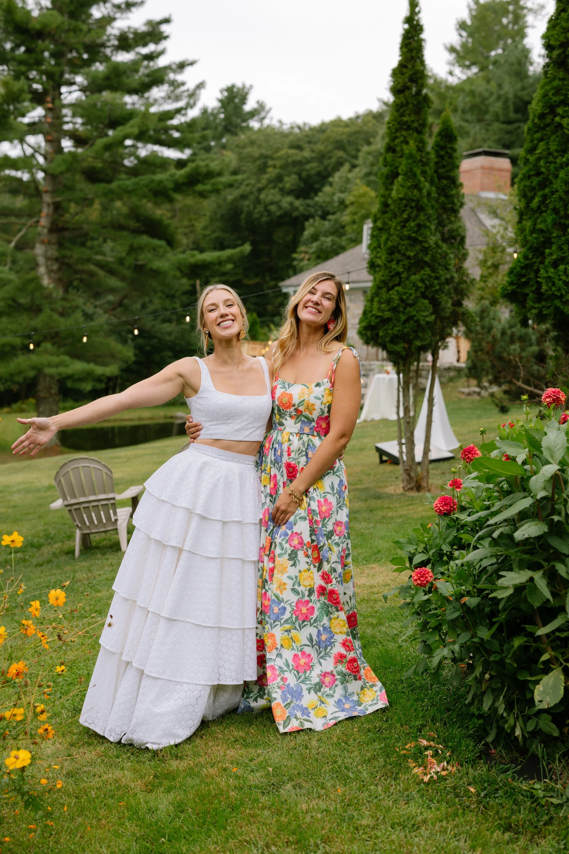 Two women smiling and posing outdoors in a garden, surrounded by trees, flowers, and a house in the background, during daytime.