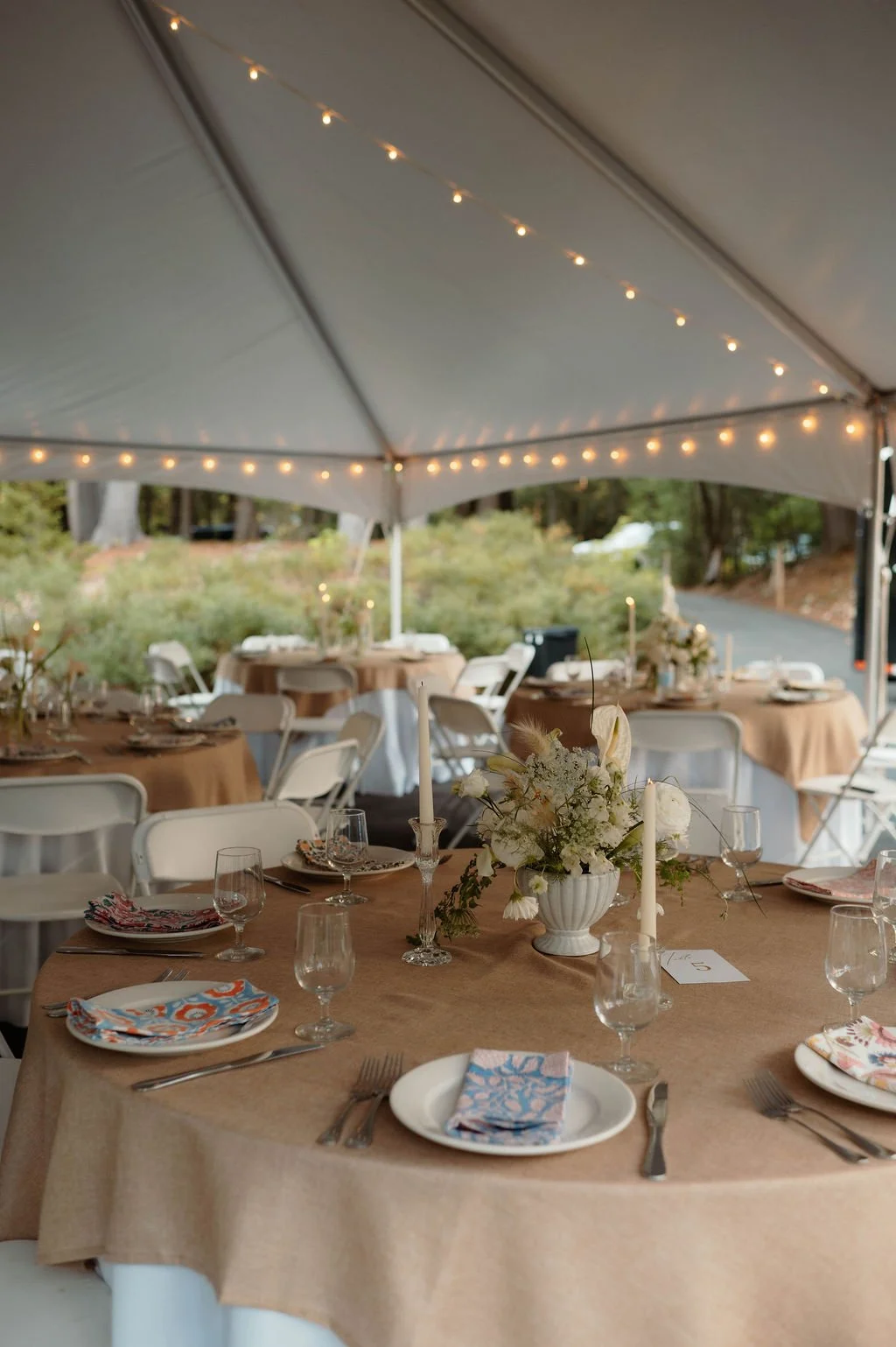 Wedding reception setup under a tent with round tables covered with beige tablecloths, floral centerpieces, candles, and place settings with glassware and napkins.
