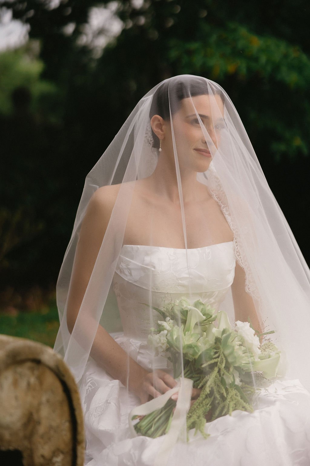 A bride in a white wedding dress with a veil holding a bouquet of white and green flowers outdoors with greenery in the background.