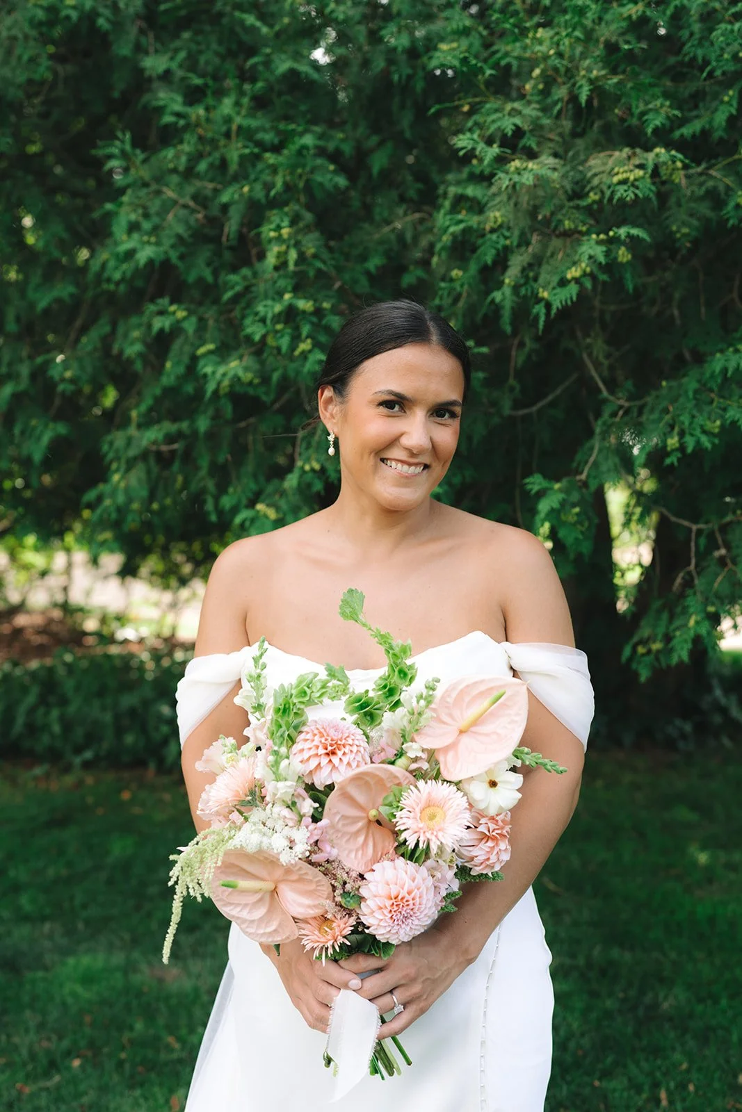 A smiling bride in a white off-shoulder wedding dress holding a bouquet of pink and white flowers standing outdoors in front of green trees.