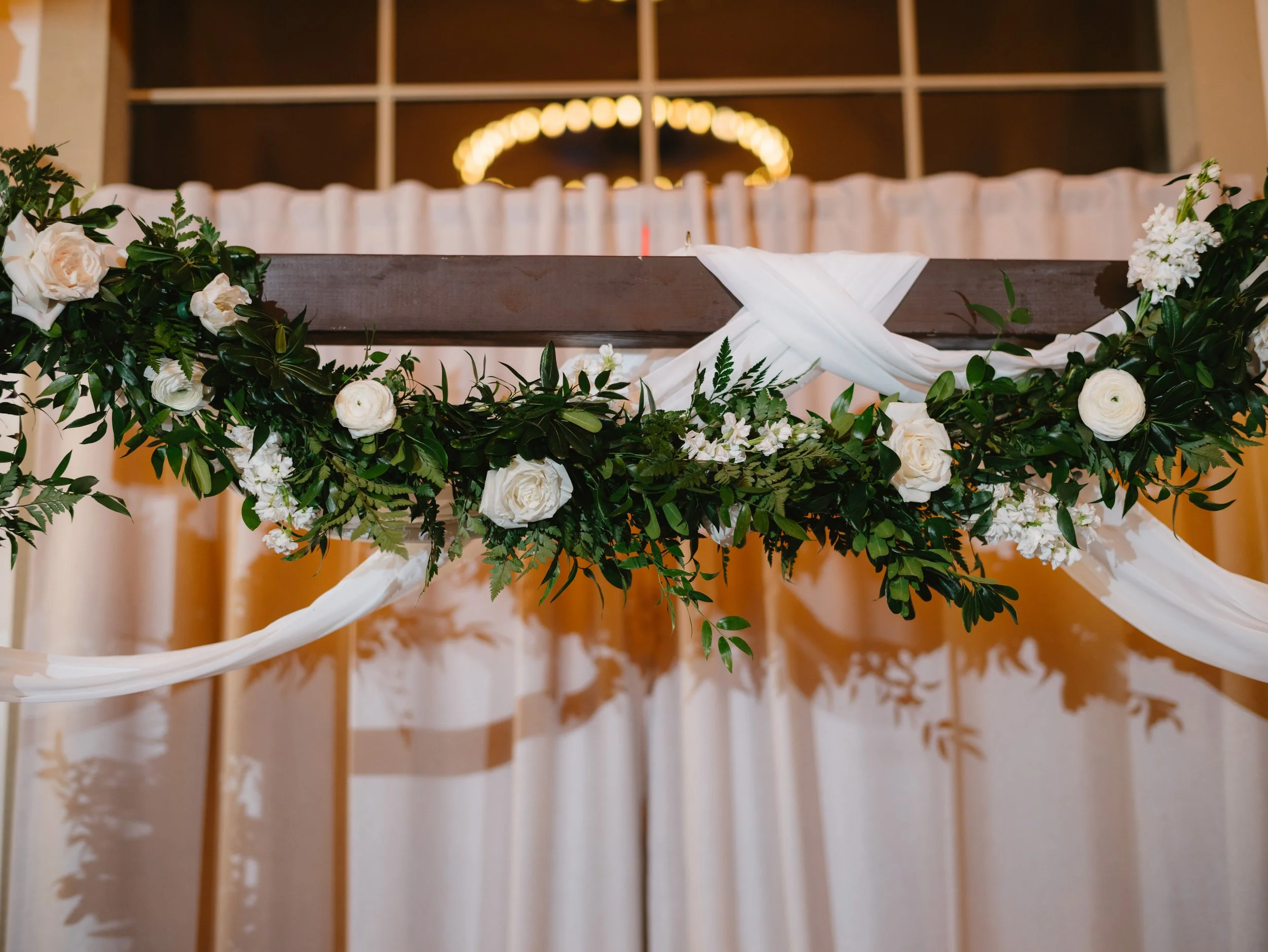 Close-up of a wedding altar decorated with white roses, greenery, and white fabric, with a backdrop of beige curtains and a lit chandelier