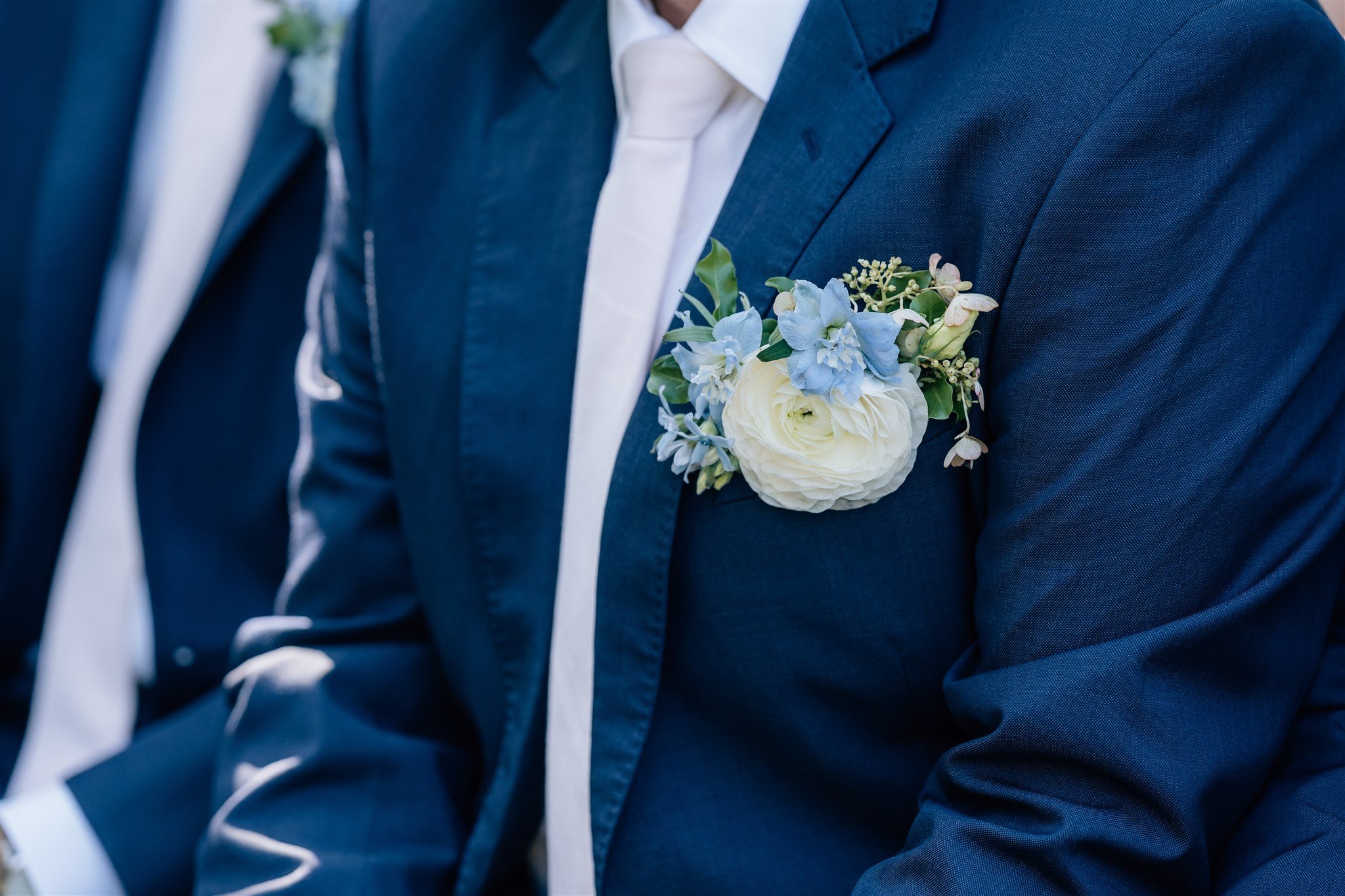 Close-up of a man in a dark blue suit with a white dress shirt, wearing a boutonniere with white and light blue flowers on his lapel at a formal event.