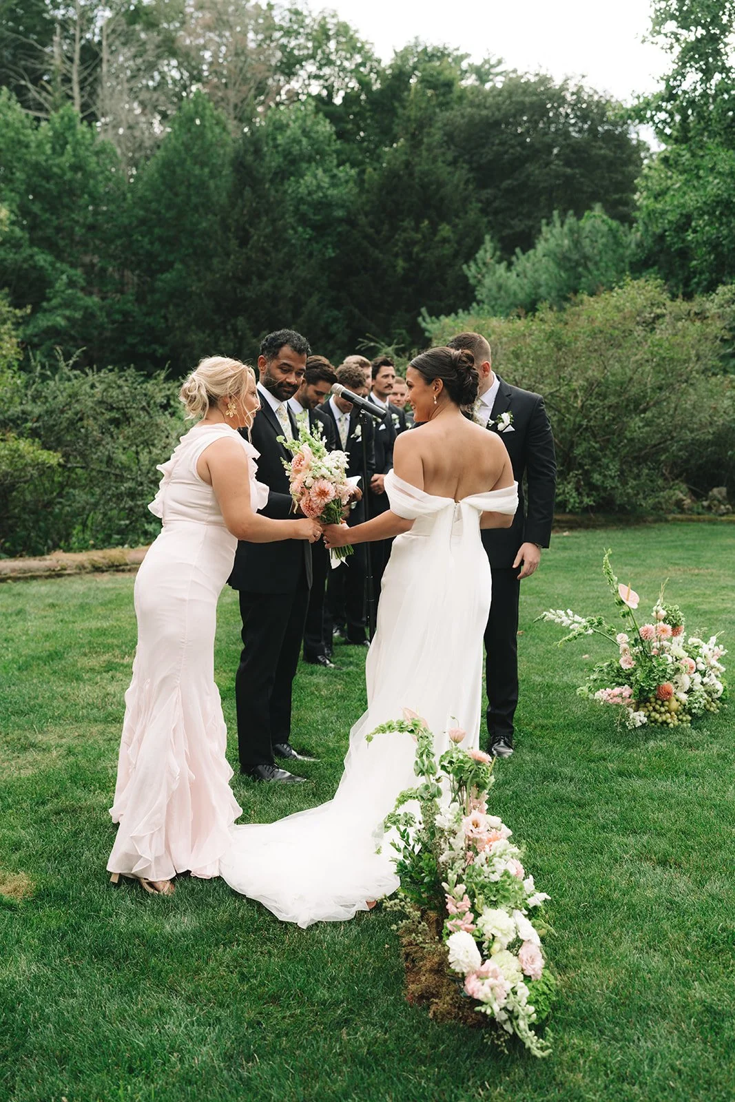 A wedding ceremony taking place outdoors on a grassy area with trees in the background. The bride, in a white off-the-shoulder gown, is holding a bouquet and facing the groom, who is in a black tuxedo. The officiant and wedding party, dressed in form