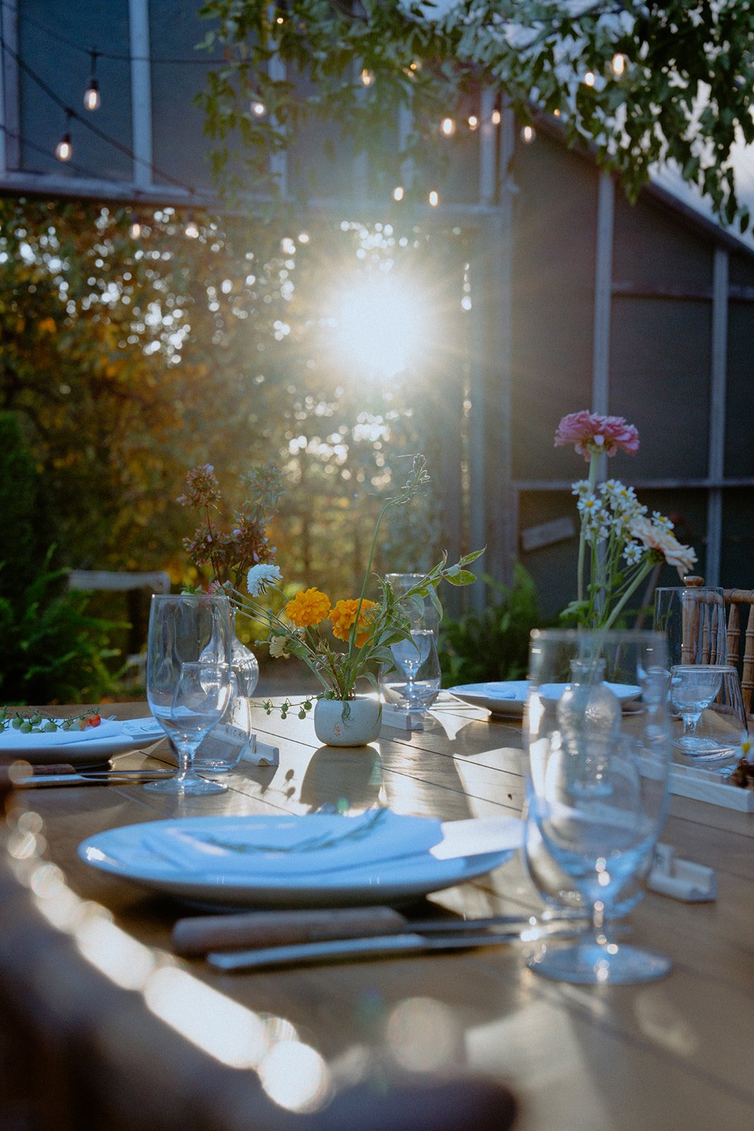 A beautifully set outdoor dining table illuminated by sunlight, decorated with flower arrangements and glassware.