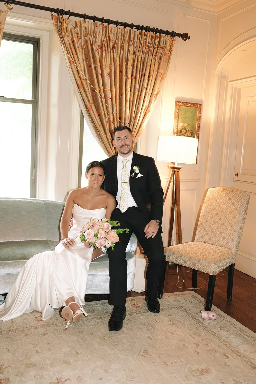A bride and groom sitting on a vintage sofa inside a well-lit room with elegant decor. The bride is holding a bouquet of pink flowers and wearing a white strapless wedding dress, while the groom is in a black suit with a floral tie. There is a window