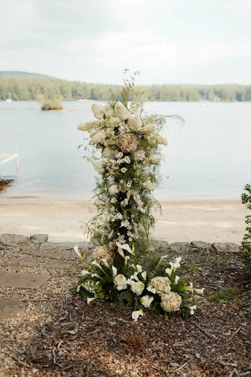 A floral arrangement with white and pale pink flowers by the water with sandy shore, trees, and sky in the background.