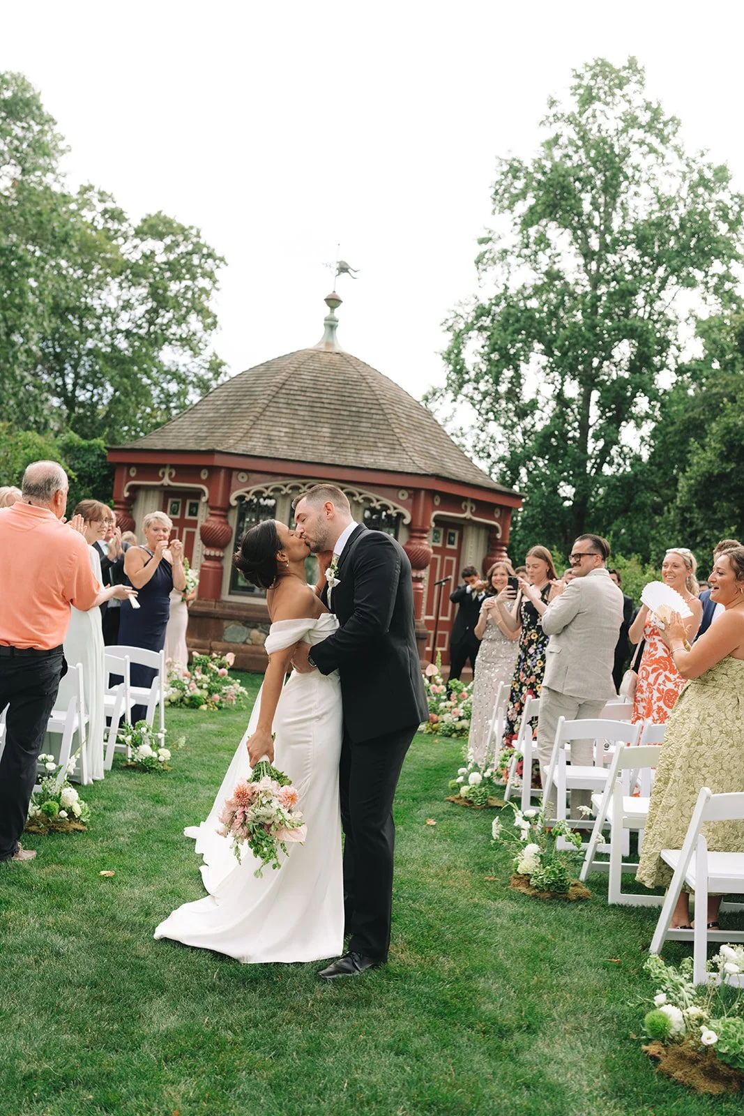 A bride and groom kiss during their outdoor wedding ceremony, surrounded by guests on a lush green lawn