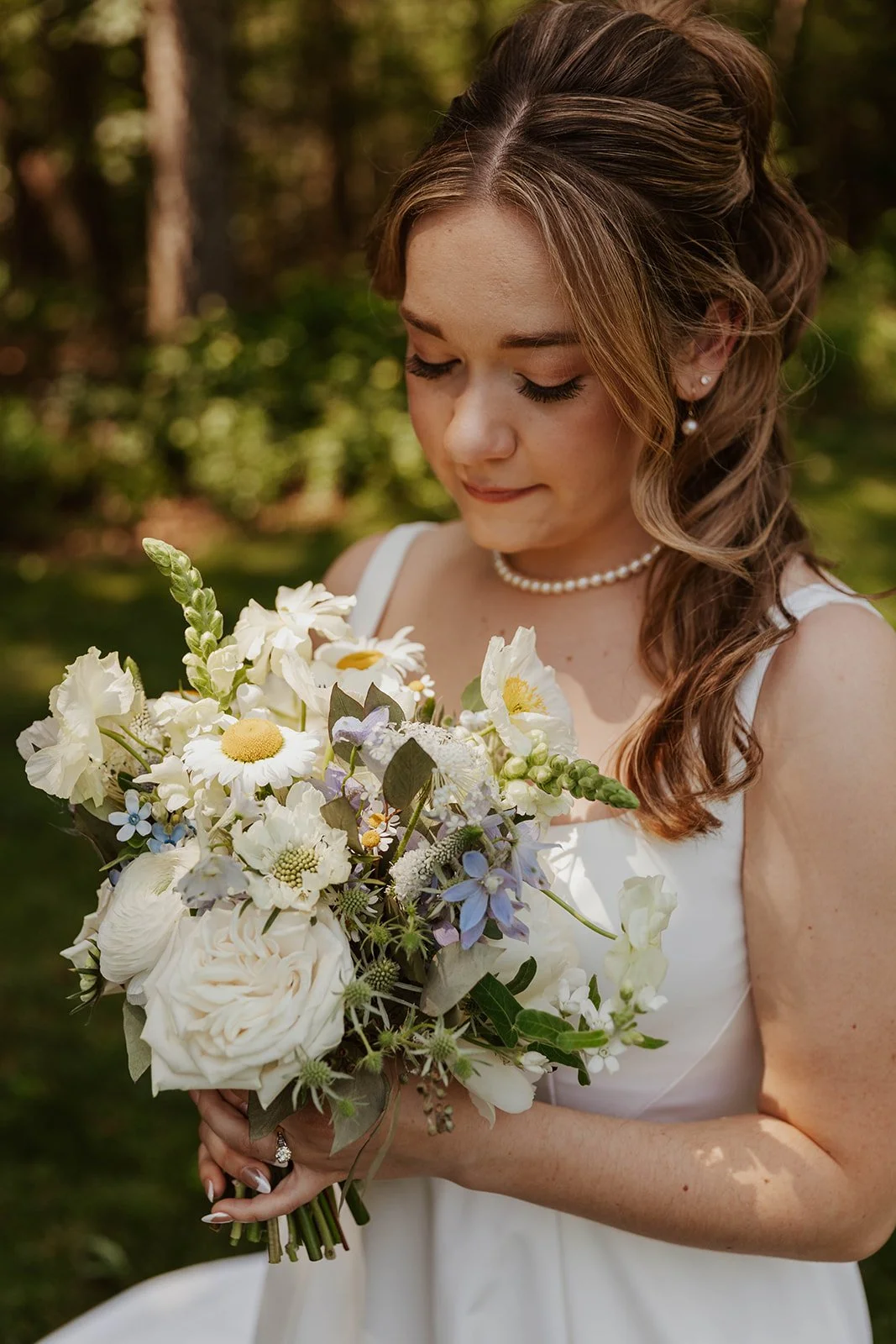 A bride holding a bouquet of white and pastel flowers, wearing a white dress, pearl necklace, and earrings, outdoors with sunlight filtering through the trees.