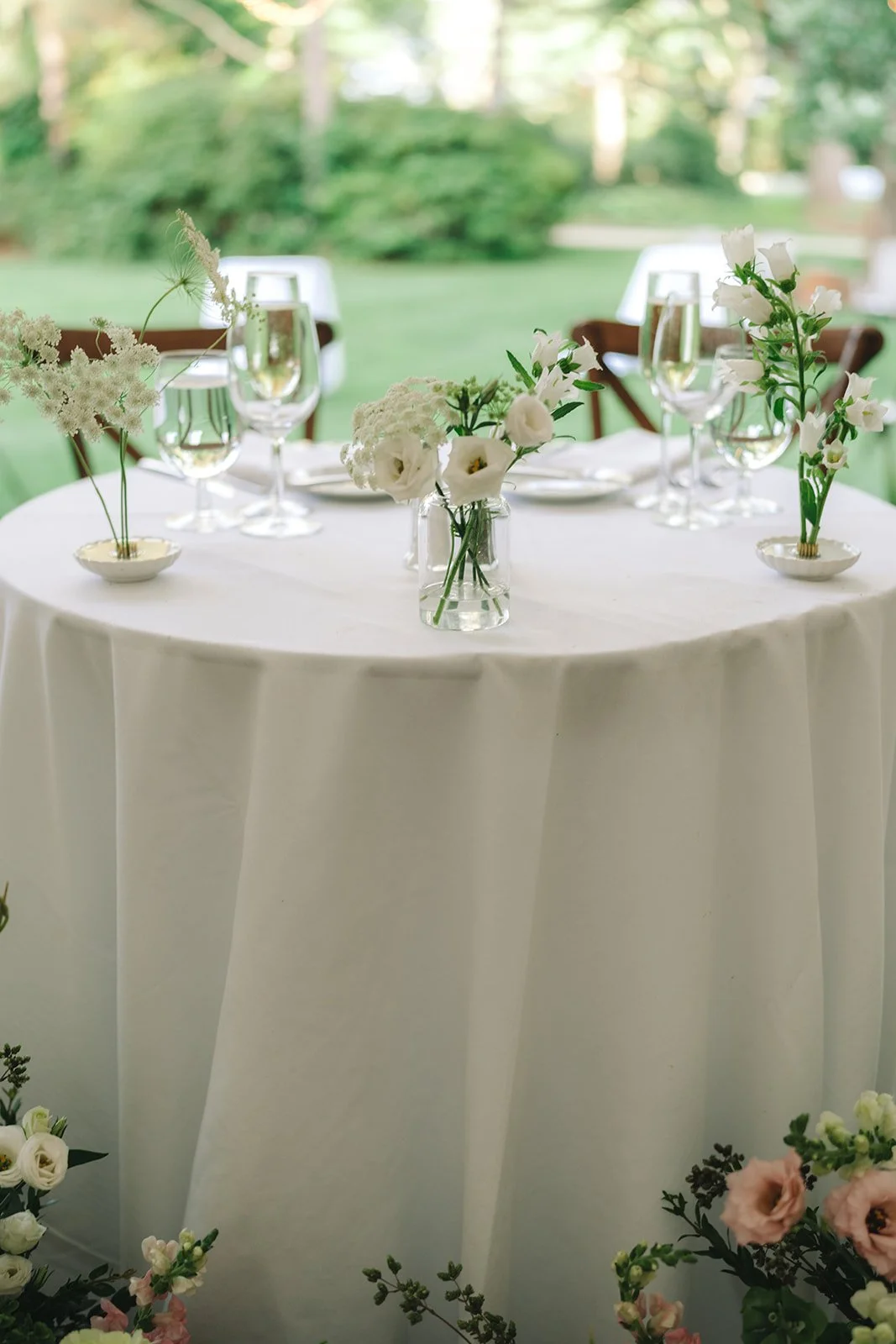 Round table with white tablecloth and floral centerpieces in vases and small bowls, set outdoors with green trees in the background.