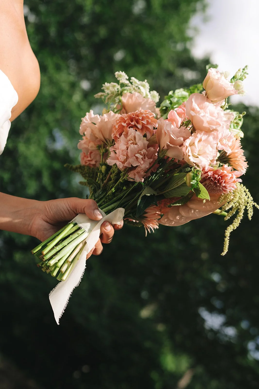A person holding a bouquet of pink and peach flowers with green stems, outdoors with blurred green trees in the background.