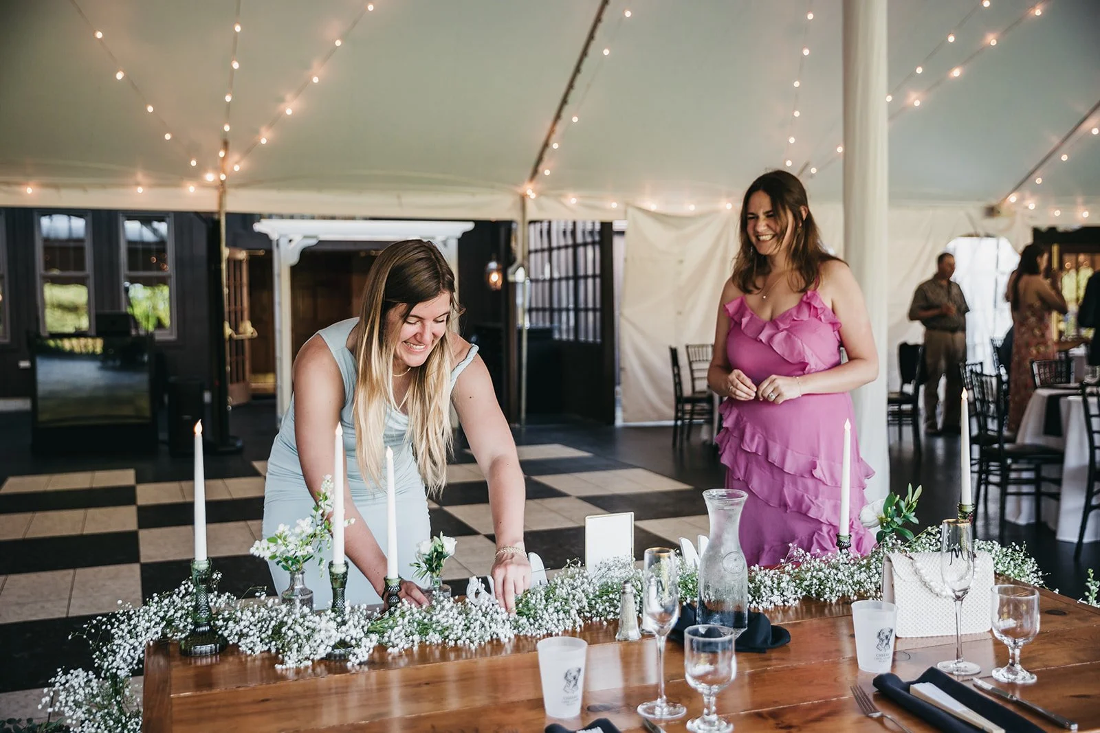 Two women in dresses are smiling and laughing at a decorated table during a celebration event. One woman in a light blue dress is leaning over the table, and the other in a pink ruffled dress is standing beside her. The table is adorned with white fl