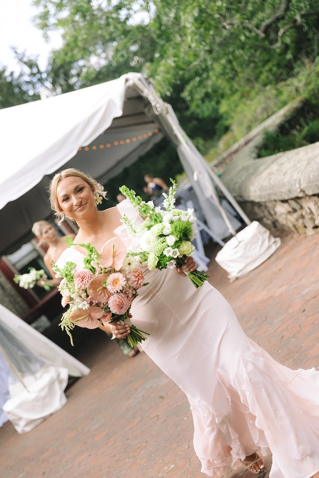 A woman in a wedding dress holding two bouquets of white and pink flowers at an outdoor wedding.