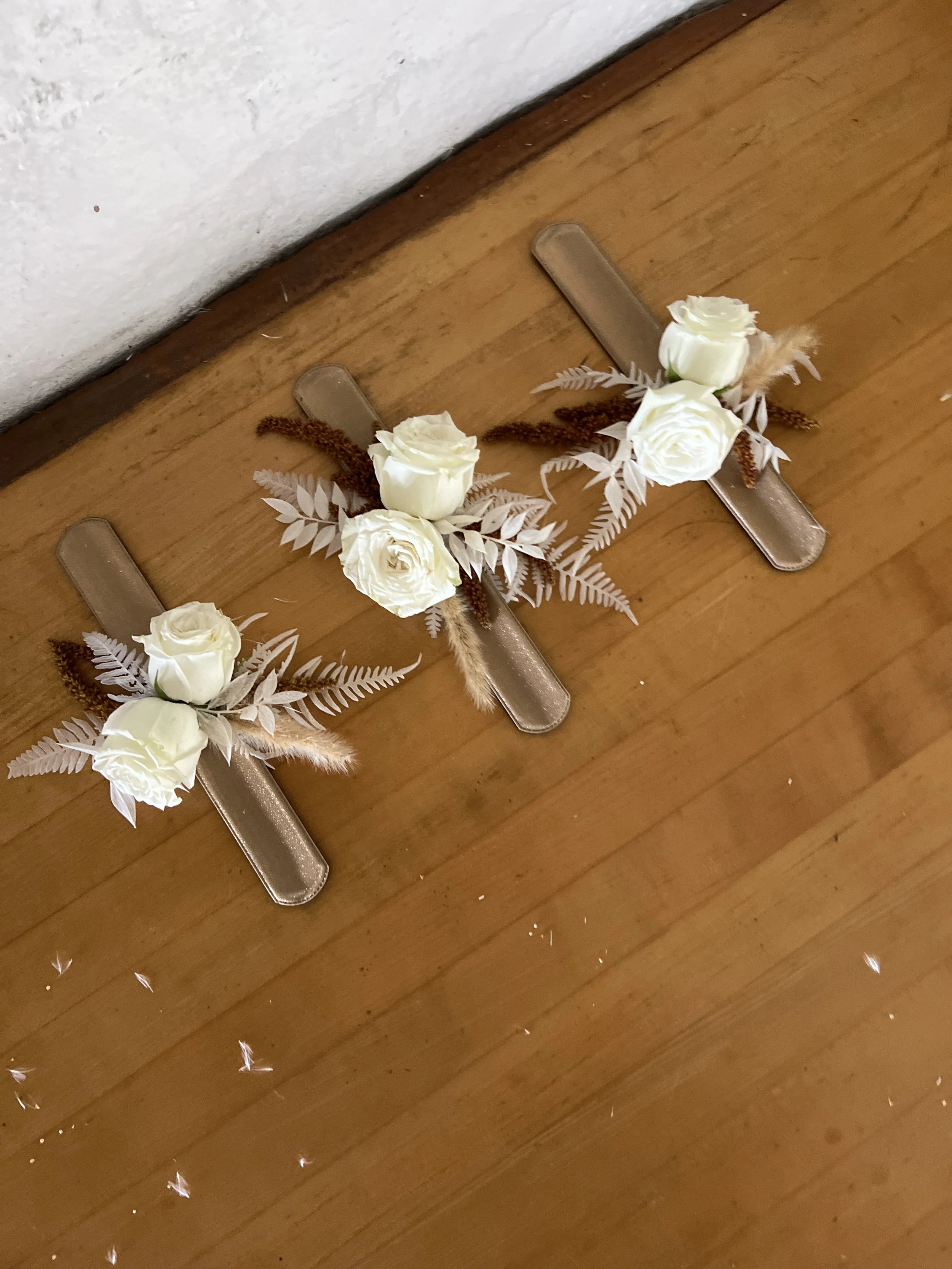 Three decorative floral pieces with white roses, beige leaves, and faux foliage arranged on golden metallic strips, placed on a wooden floor near a white wall.