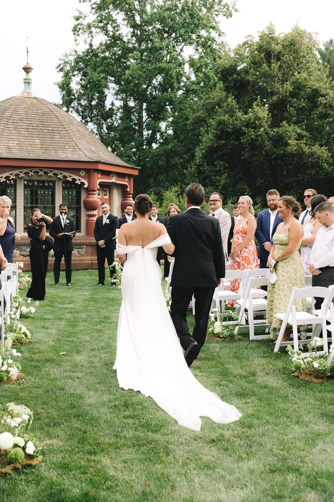A bride and groom walking down the outdoor wedding aisle. The bride is in a white off-shoulder wedding gown, and the groom is in a black suit. Guests are seated on either side, watching and smiling. The wedding is in a garden with trees and a gazebo 