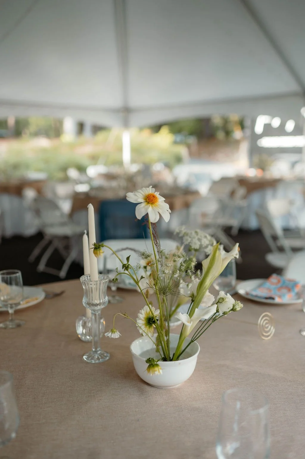 A floral centerpiece with white and yellow flowers on a tan tablecloth at an outdoor event under a large white tent.