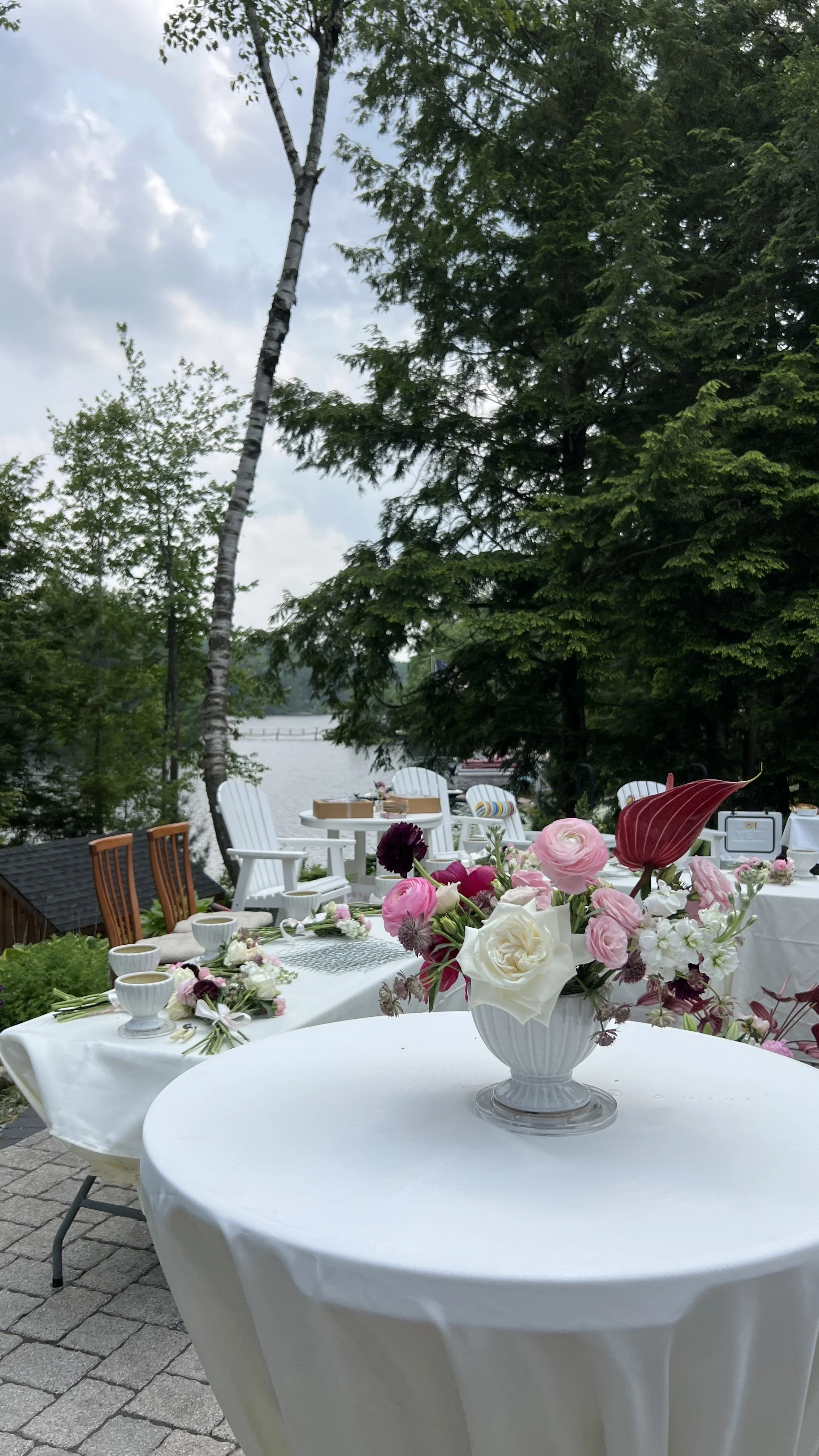Outdoor setting with white tables and chairs, decorated with pink and white flowers, overlooking a lake with trees surrounding the area.