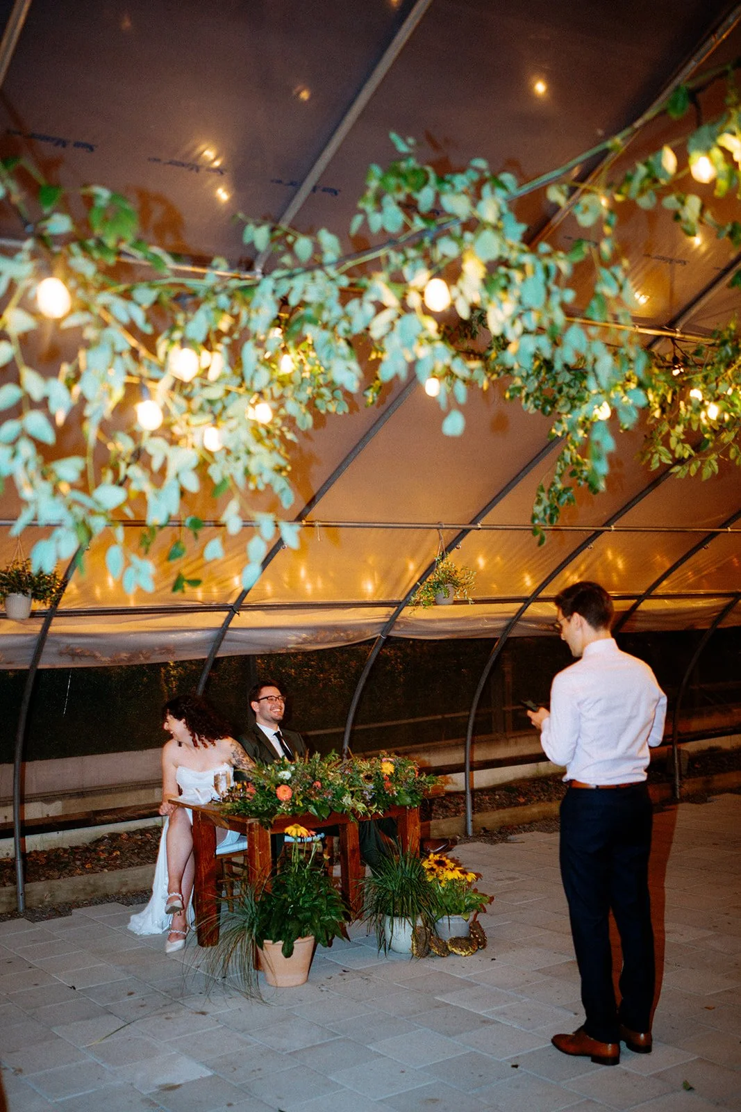 A wedding ceremony takes place under a canopy decorated with greenery and string lights. The bride and groom sit at a small table adorned with flowers, while a man, possibly a guest or officiant, stands nearby using his phone.
