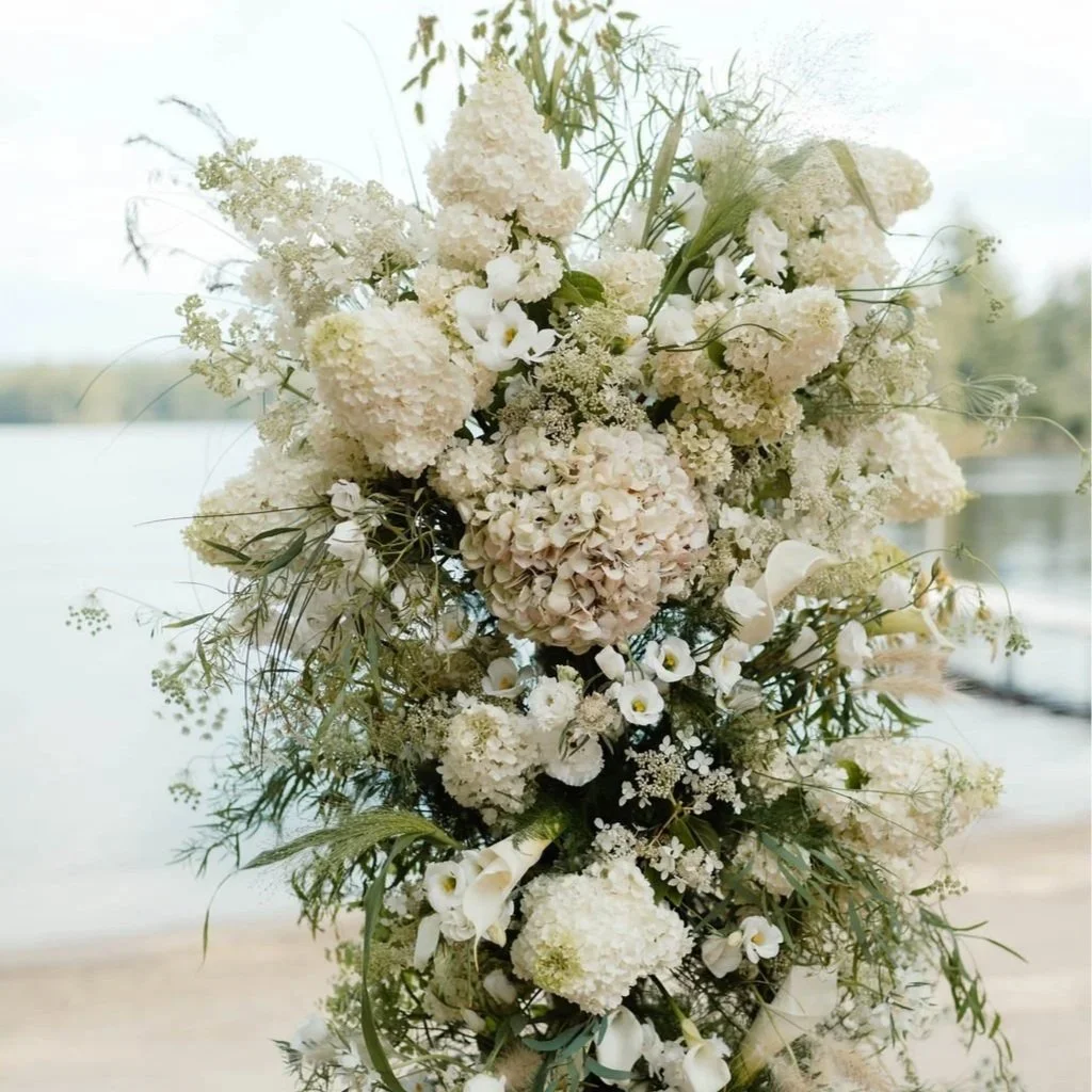 A tall, cascading bouquet of white and cream flowers, including hydrangeas, calla lilies, and small filler blooms with greenery, set against a blurred outdoor background.
