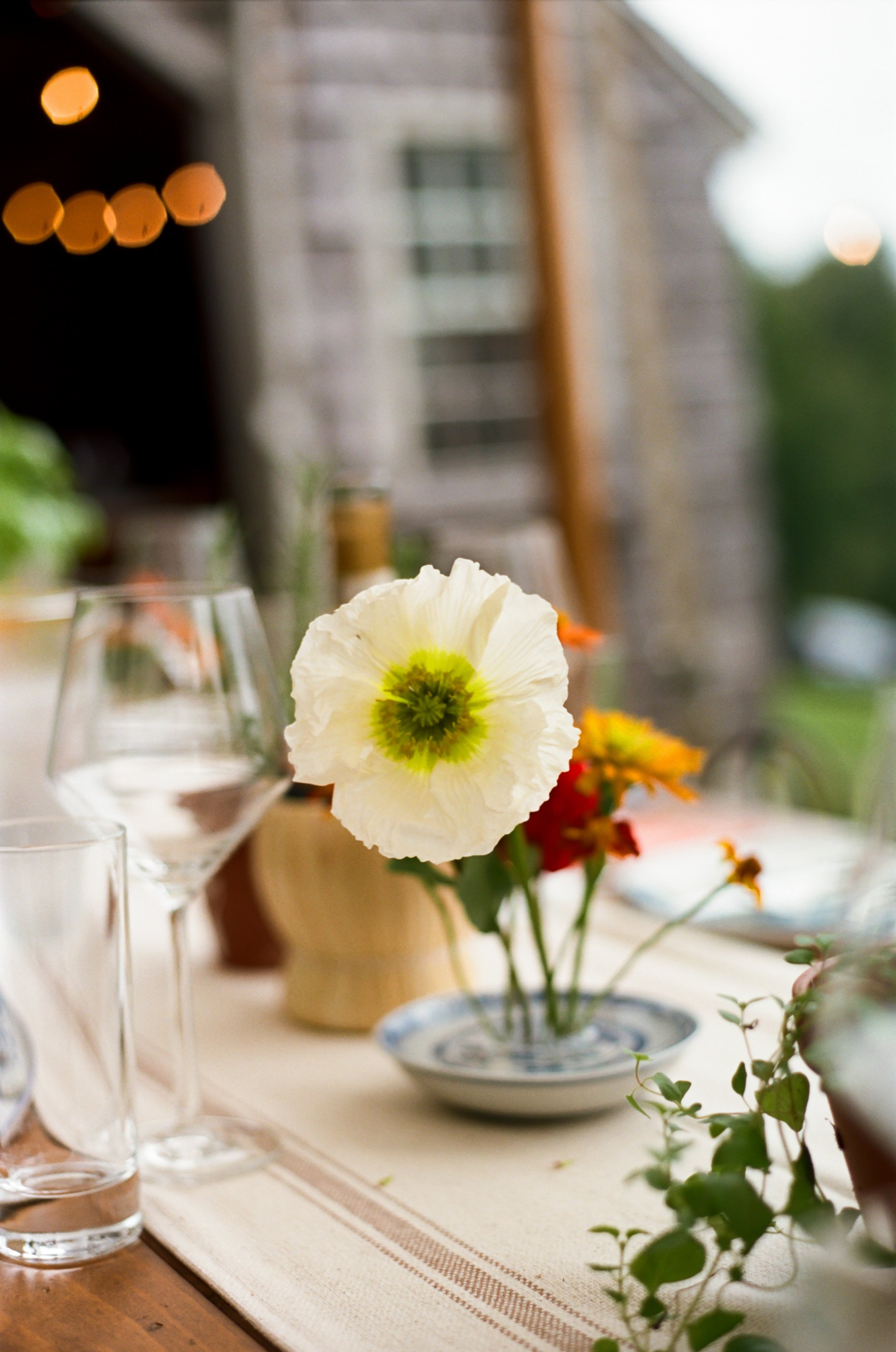 Close-up of a white flower with a green center in a small bowl on a dining table, surrounded by empty glasses and other flowers, with a blurred outdoor background.