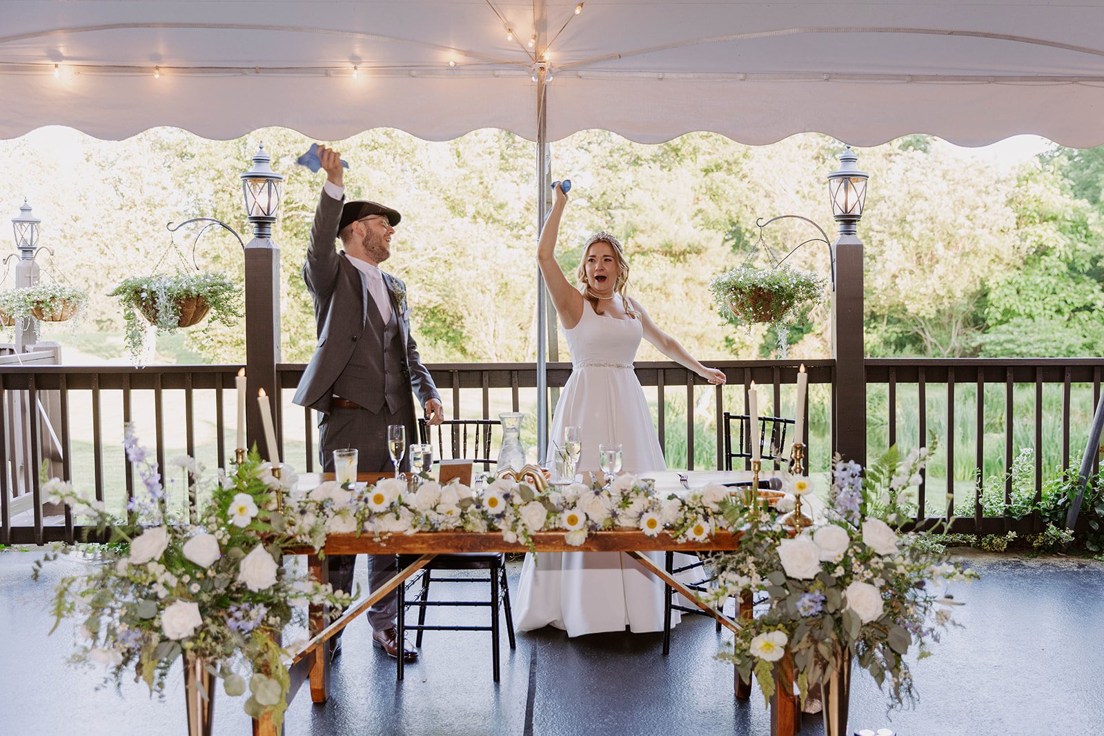 A bride and groom celebrate at their wedding reception, raising glasses in a toast under a large white canopy decorated with string lights, with trees in the background and floral arrangements on the table.