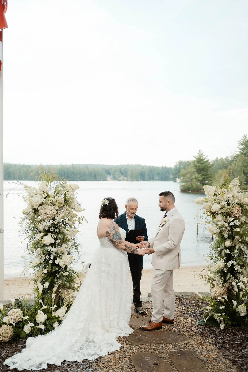 A wedding ceremony taking place outdoors by a lake, with a couple holding hands and an officiant reading from a book, surrounded by floral decorations.