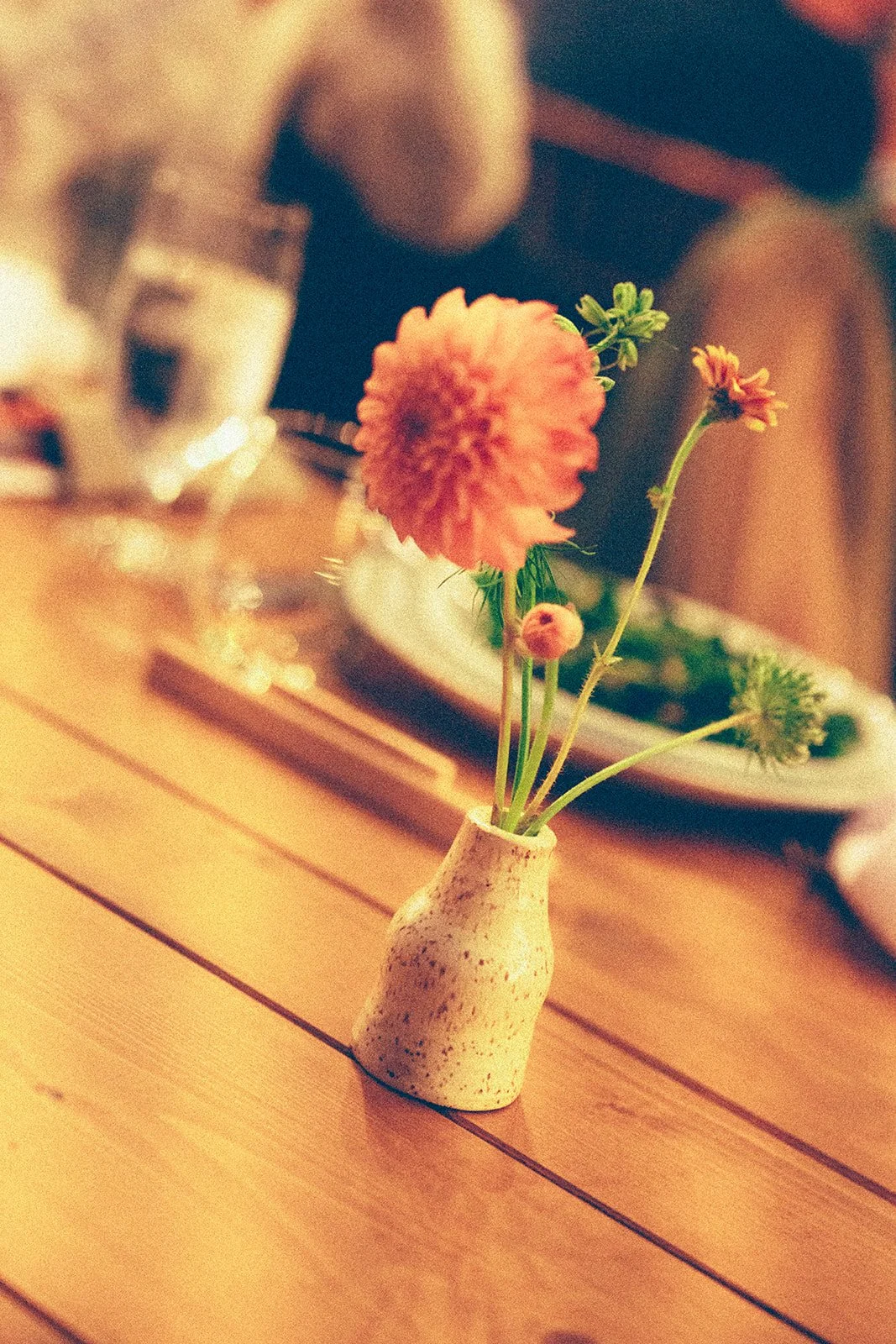 A small ceramic vase holding a few flowers, including a large, pink-dyed flower, on a wooden table in a cozy restaurant setting.