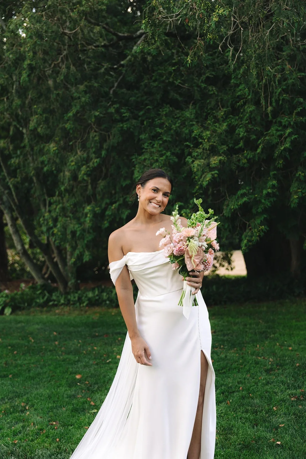 A woman in a white off-the-shoulder wedding dress holding a bouquet of pink and white flowers outdoors with greenery in the background.