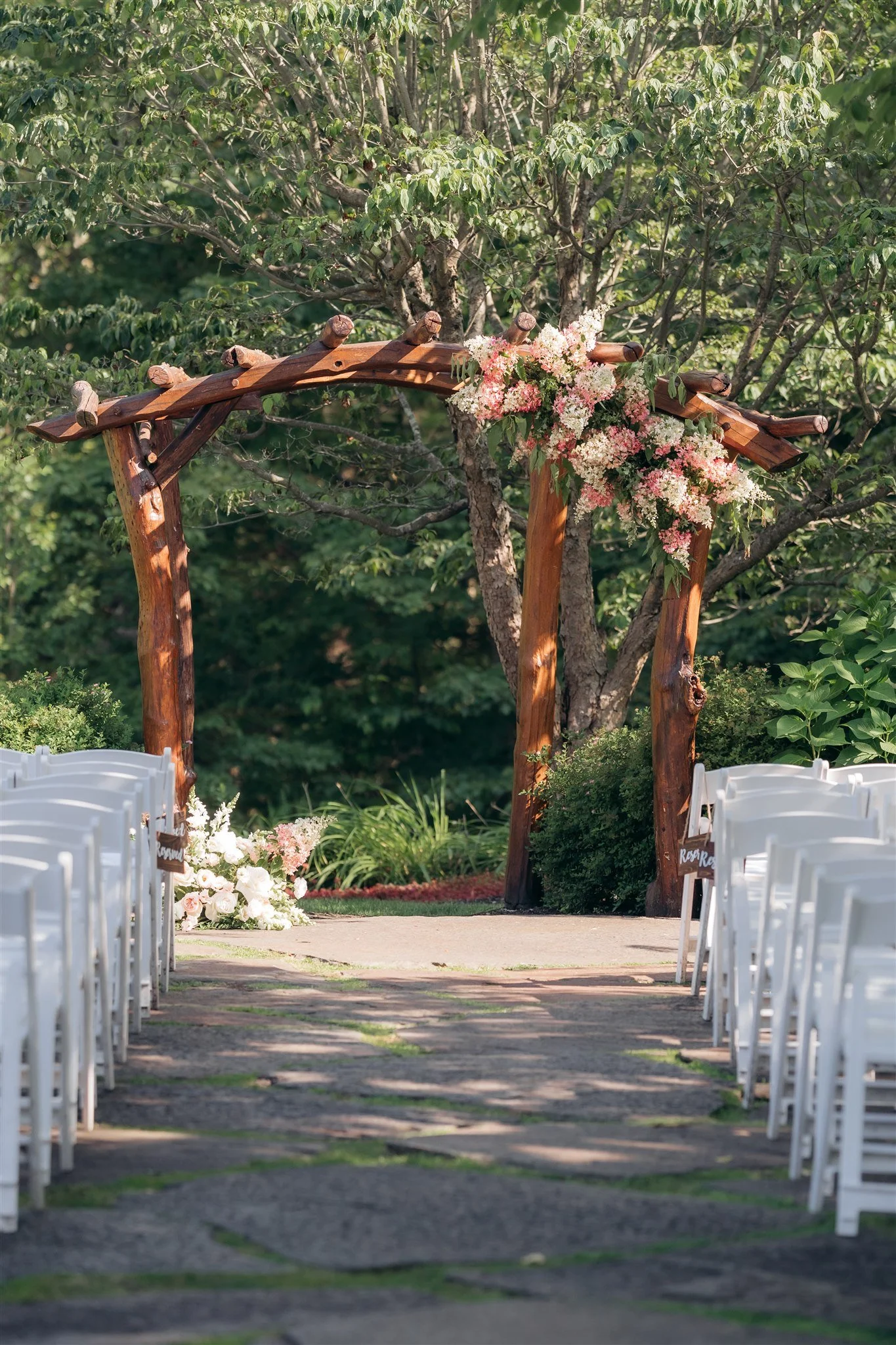 Wedding ceremony setup with white chairs on either side of an aisle, leading to a wooden arch decorated with pink and white flowers, in an outdoor garden with lush green trees.