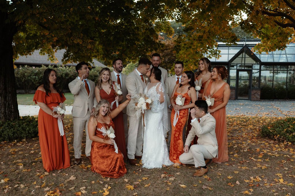Group of wedding guests, with the bride and groom, celebrating outdoors under a large tree with fall foliage behind them.