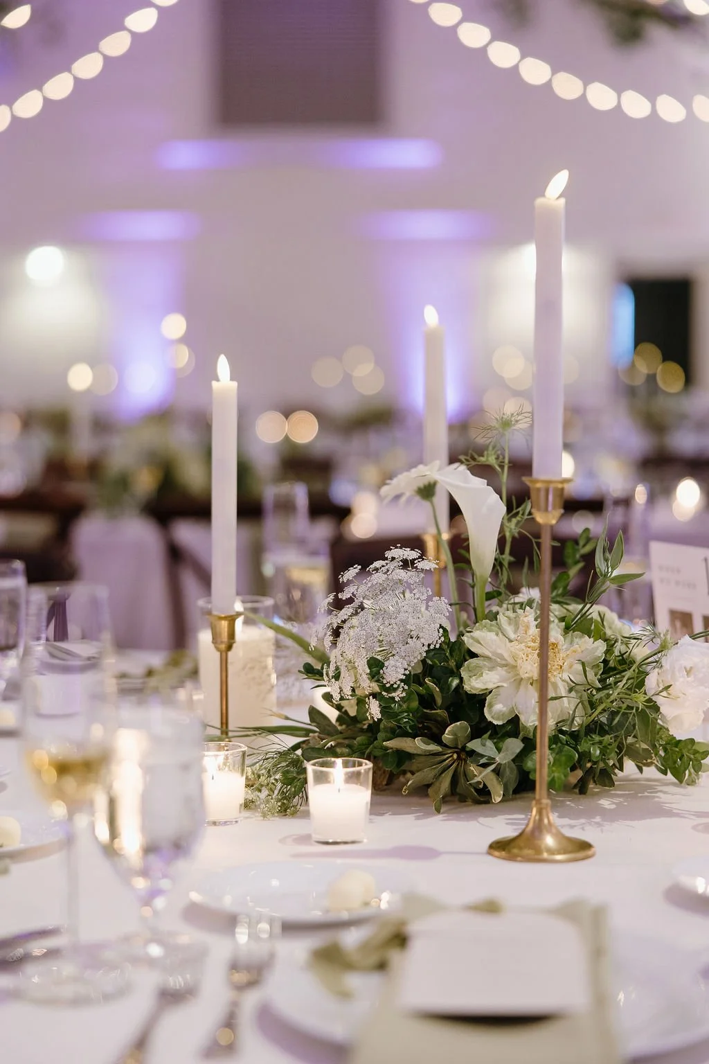 Elegant wedding reception table with tall white candles, floral centerpiece with white flowers and greenery, lit candles, glassware, and place settings.