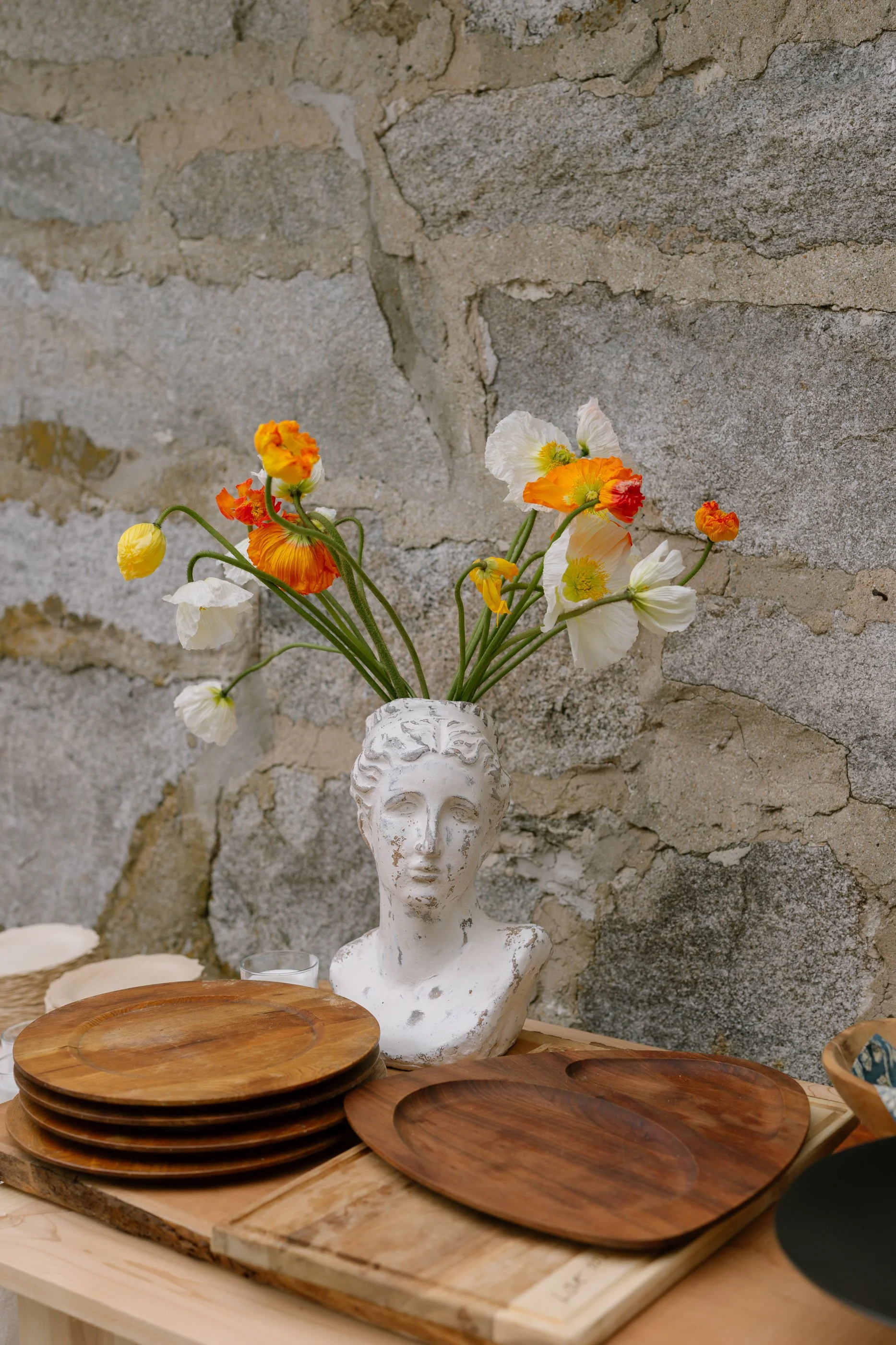 A decorative display featuring a white bust-shaped vase with colorful poppy flowers on a wooden table, set against a stone wall background.