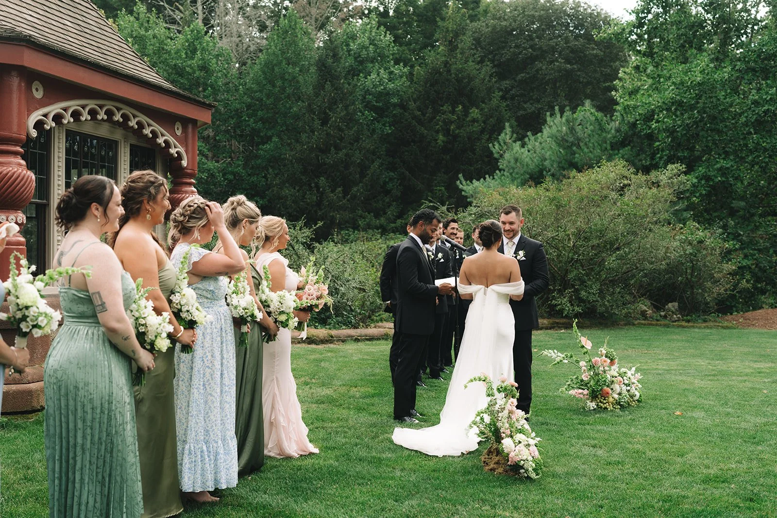 Outdoor wedding ceremony with a bride and groom exchanging vows, surrounded by bridesmaids holding bouquets and standing outdoors on a grassy lawn with lush greenery in the background.