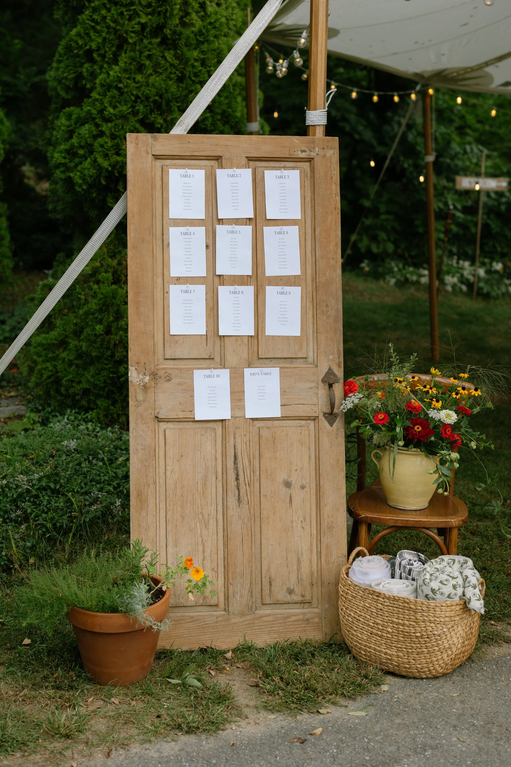 A wooden outdoor seating chart board with white paper cards for each table, labeled from 1 to 10 and kids' table, set up at a garden event with flowers and potted plants nearby.