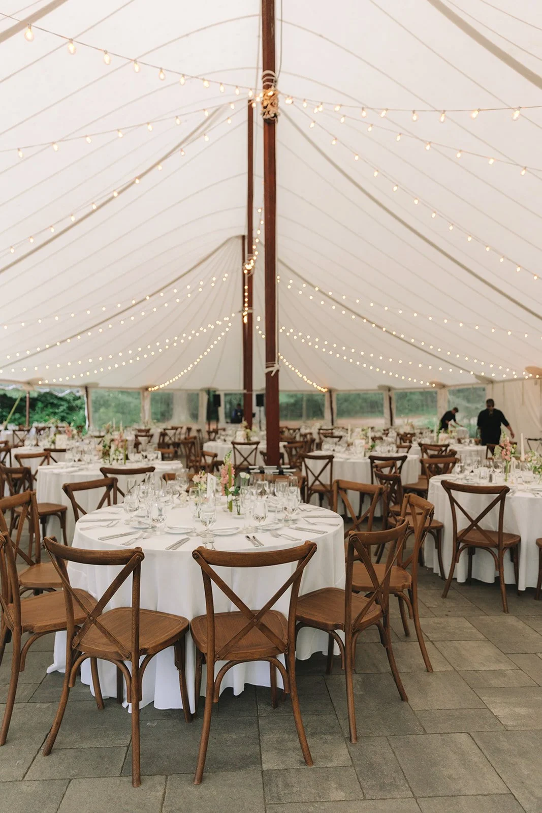 Elegant event tent with round tables set for a celebration, decorated with white tablecloths, floral centerpieces, wine glasses, and dinnerware, illuminated by string lights hanging from the ceiling.
