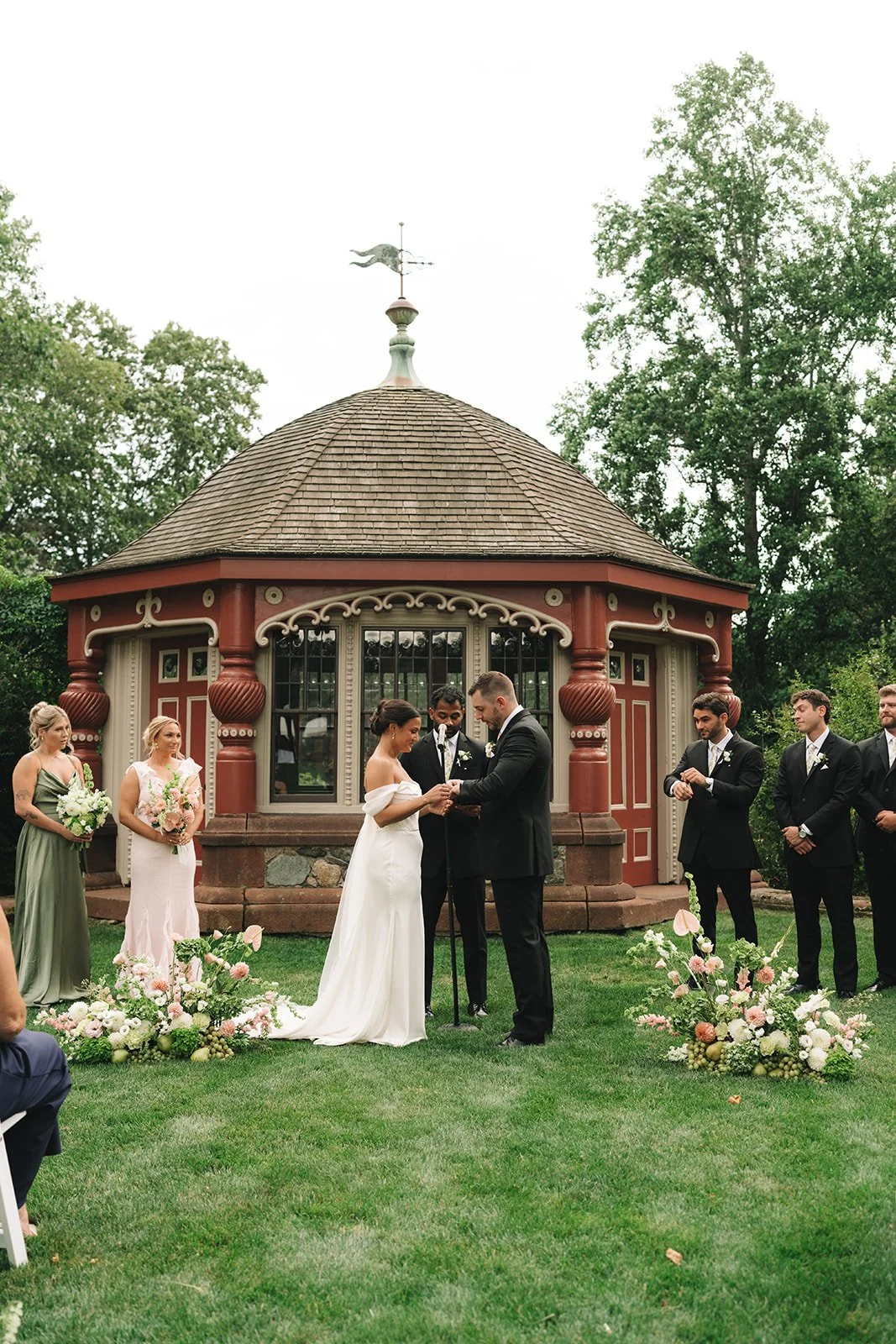 A wedding ceremony taking place outdoors in front of a small, ornate gazebo with a weather vane on top. The bride and groom are standing in the center, holding hands and exchanging vows, surrounded by bridesmaids and groomsmen.